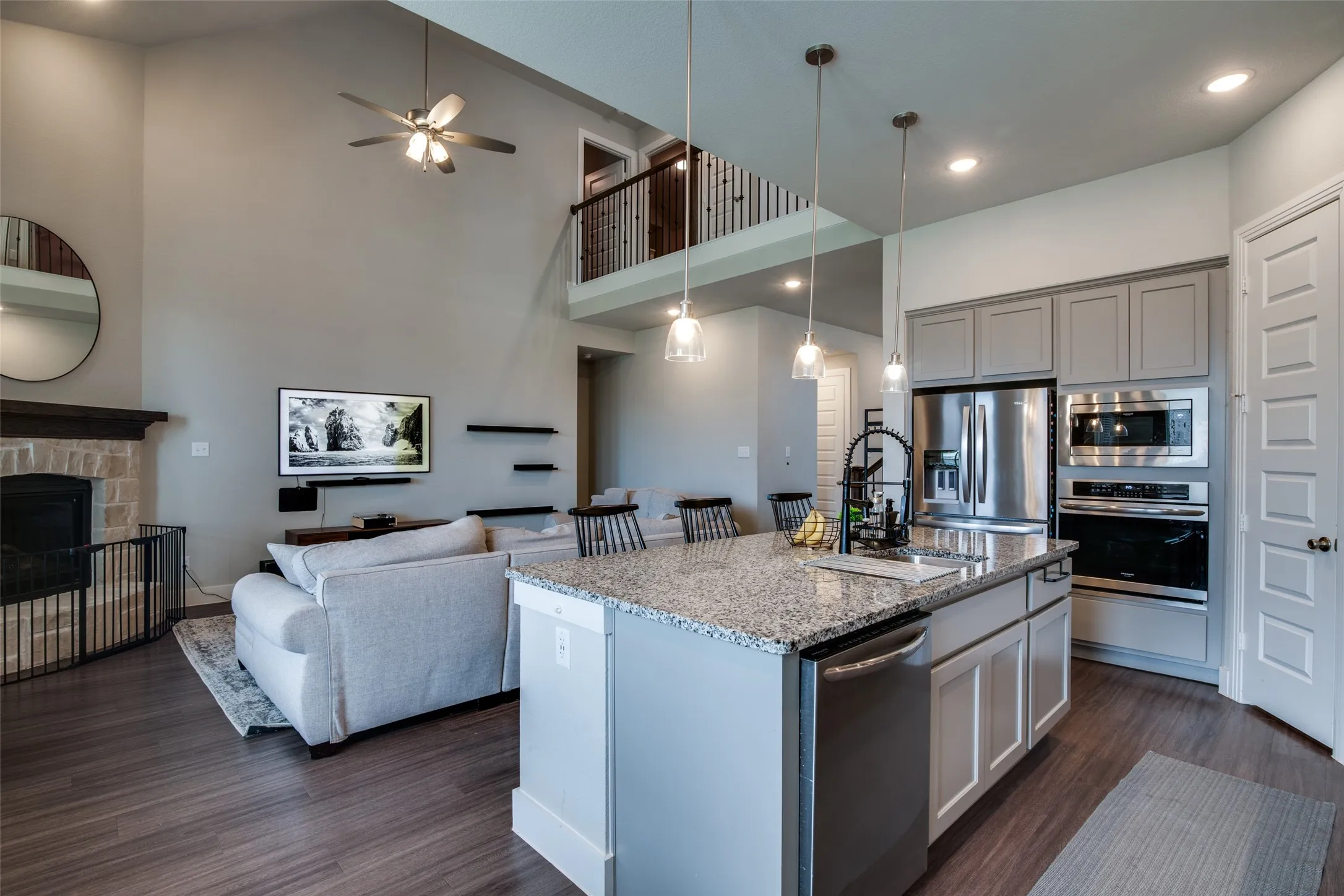 Open-concept living area featuring a kitchen island with granite-finish countertop, stainless steel appliances, wood-finish flooring, a stone fireplace, and a second-story interior balcony