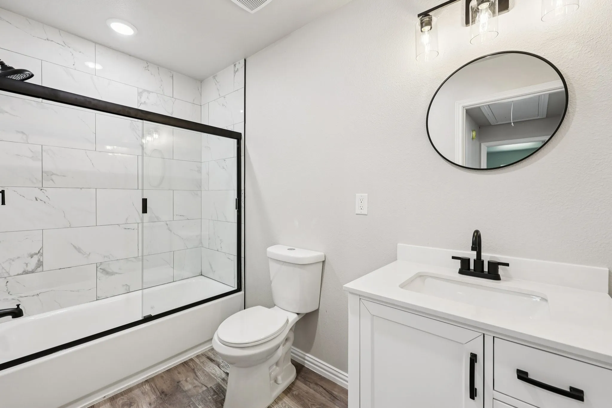 Bathroom featuring a white vanity with integrated sink, a circular mirror, and a tub with a sliding glass door enclosure