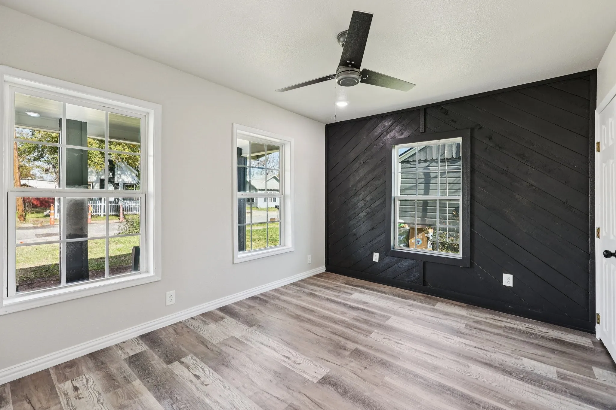 Interior space featuring a black accent wall with diagonal wood-finish paneling, three white-framed windows, wood-finish flooring, and a ceiling fan with black blades