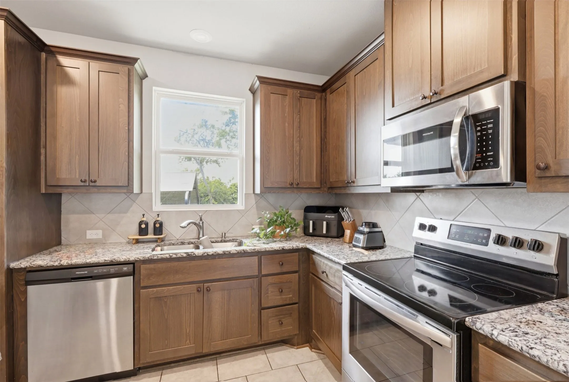 Kitchen featuring wood-finish cabinetry, granite-style countertops, stainless steel appliances, a tile backsplash with a diamond pattern, and a window above the sink