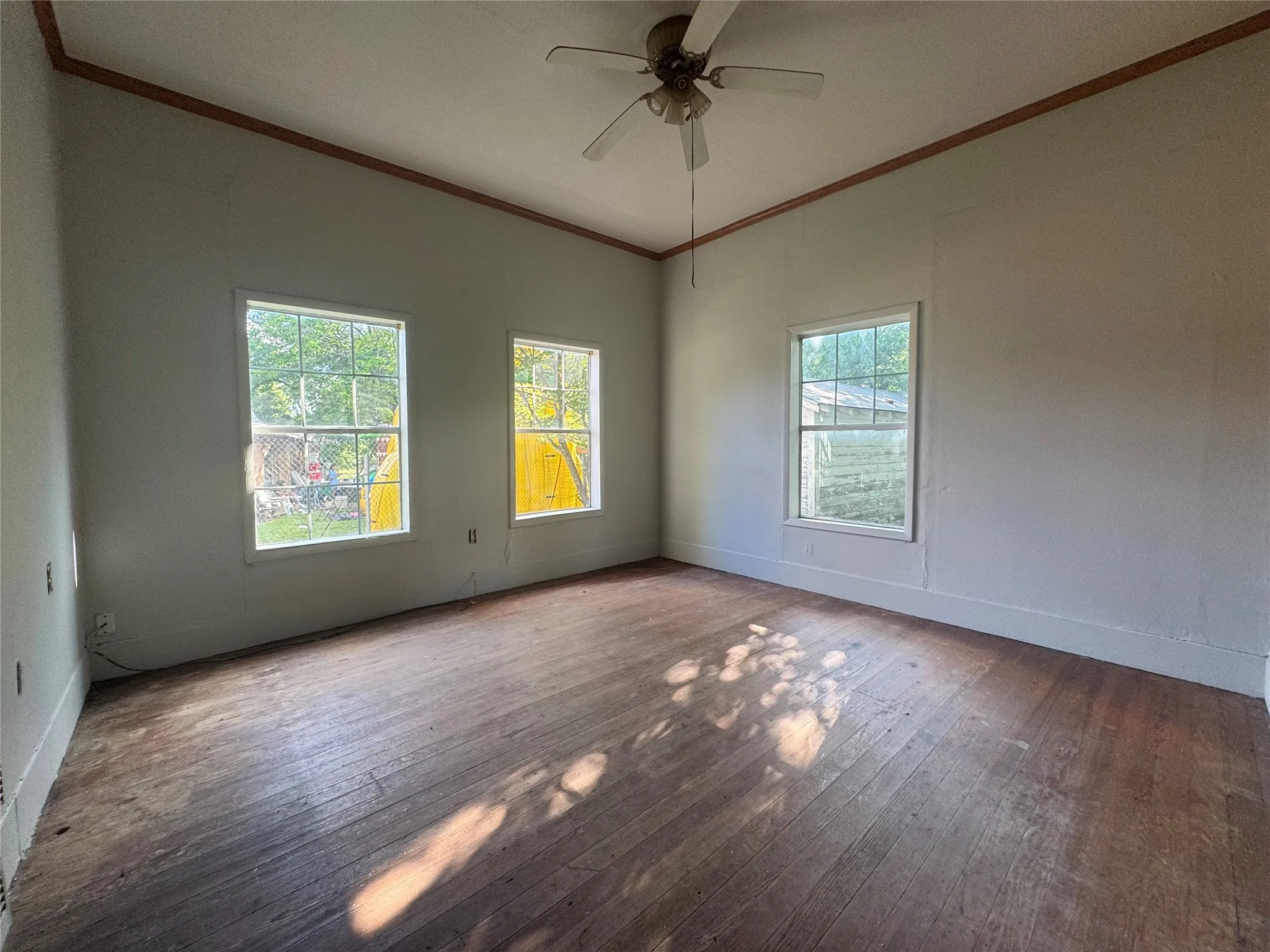Spacious room featuring three windows, wood-finish flooring, a ceiling fan, and crown molding