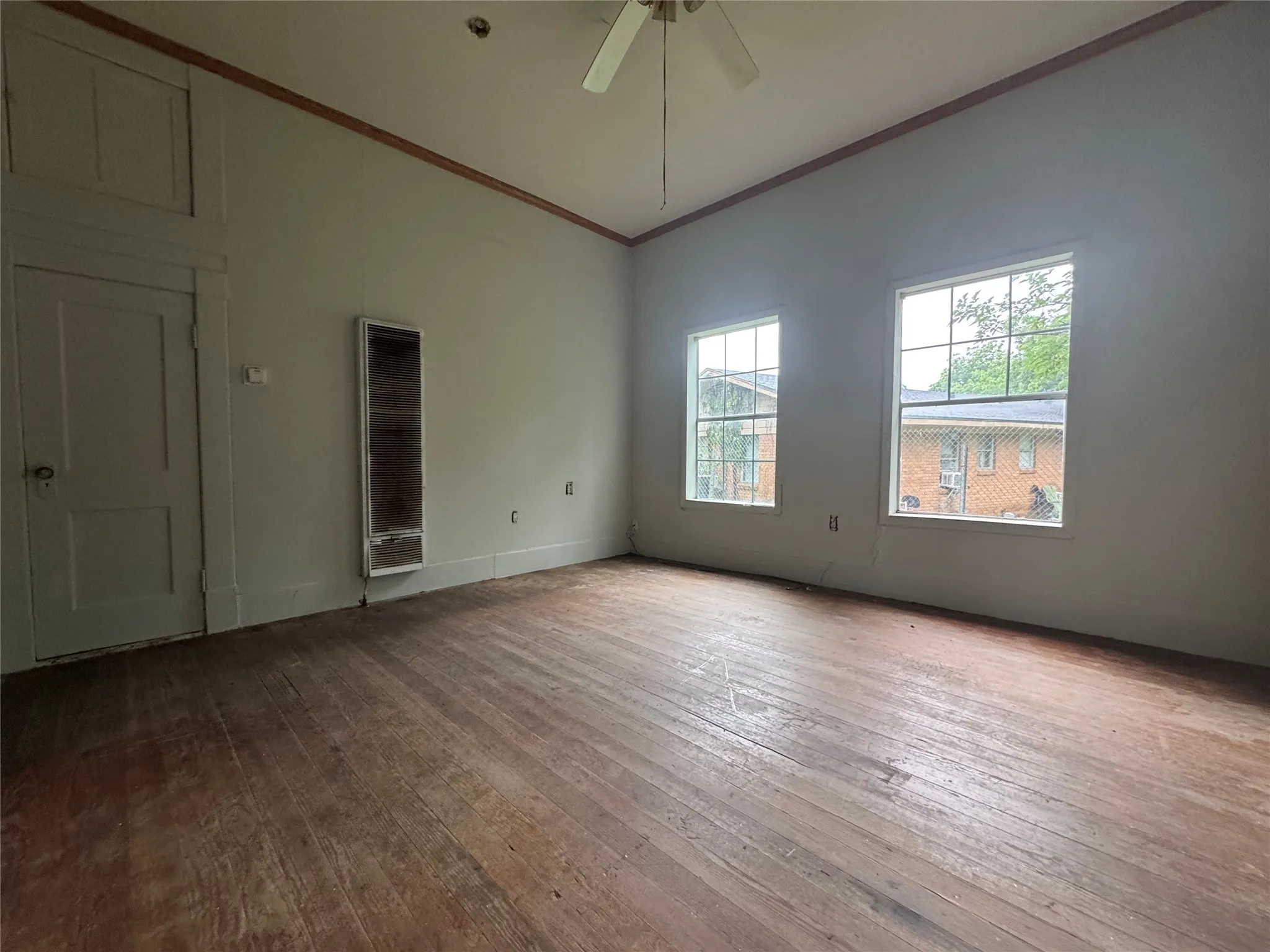 Spacious room featuring wood-finish flooring, two large windows, a ceiling fan, and a wall-mounted heating unit