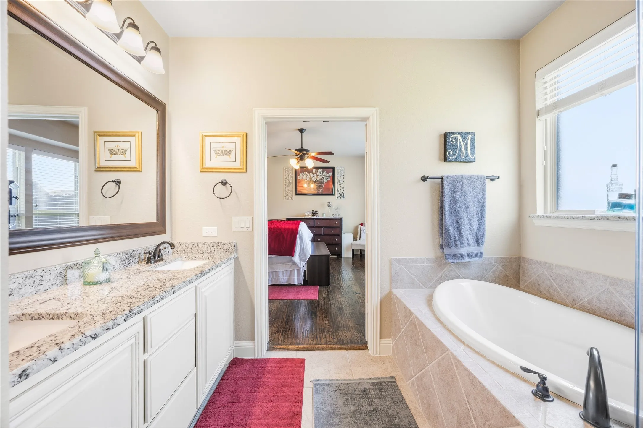 Dual sink vanity with stone countertop, mounted mirror, and built-in soaking tub with tiled surround