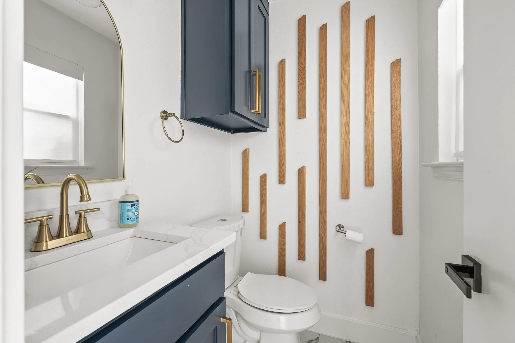 Modern powder room featuring a white single vanity with a quartz countertop, a brass-finish faucet, a rectangular mirror with a brass-finish frame, a dark blue wall-mounted cabinet, and a decorative wood wall accent