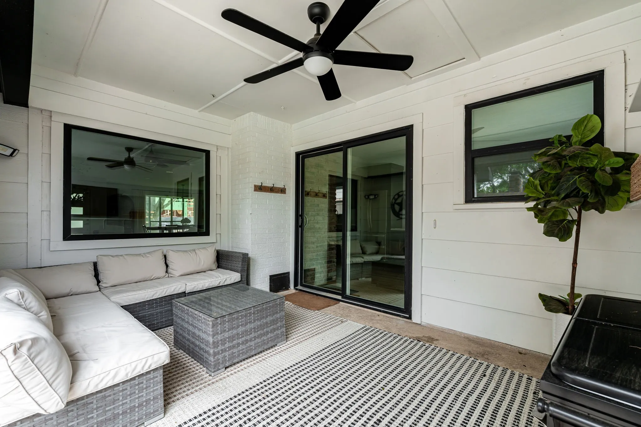 Covered patio featuring a ceiling fan, white shiplap walls, a white painted brick column, black-framed windows, and a black-framed sliding glass door