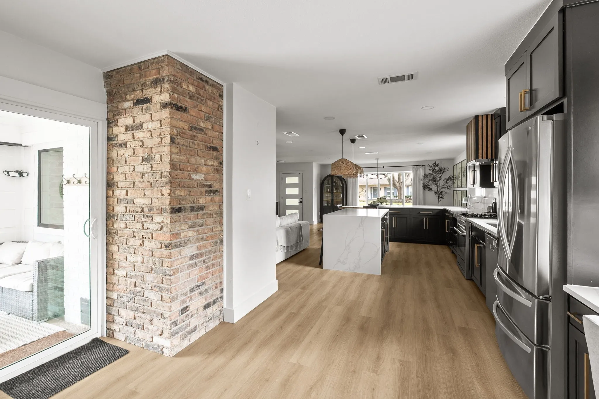 Open-concept kitchen and living area featuring wood-finish flooring, a brick accent wall, a large kitchen island with a waterfall countertop, dark cabinetry, and stainless steel appliances