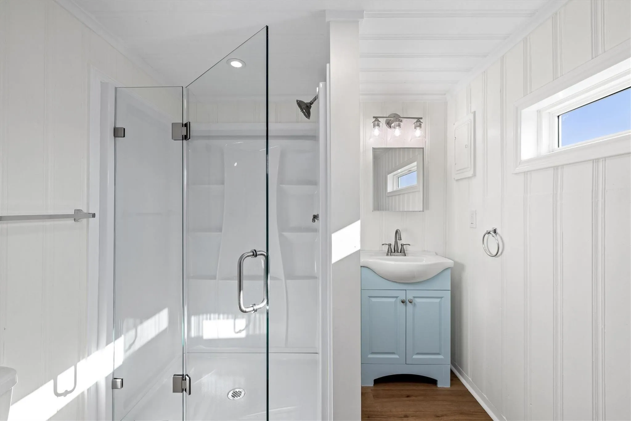 Bathroom featuring a glass-enclosed shower, a light blue vanity with an integrated sink, a vanity mirror with a three-light fixture, and wood-finish flooring