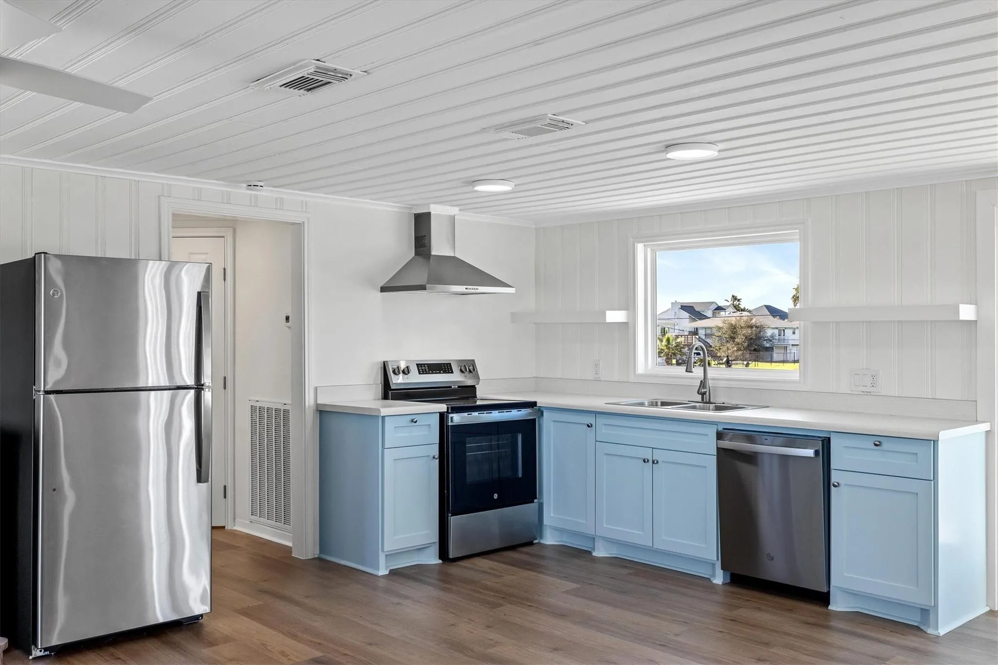Kitchen featuring light blue cabinetry, stainless steel appliances, wood-finish flooring, white paneling, and a large window above the sink