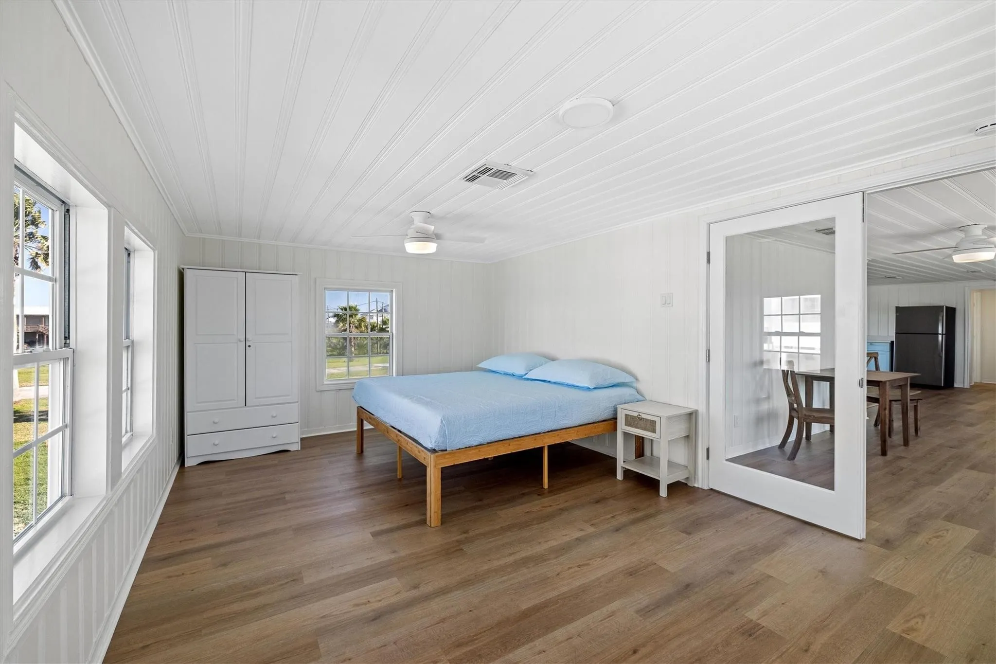 Inviting room featuring a ceiling fan, multiple windows, wood-finish flooring, and white paneled walls and ceiling