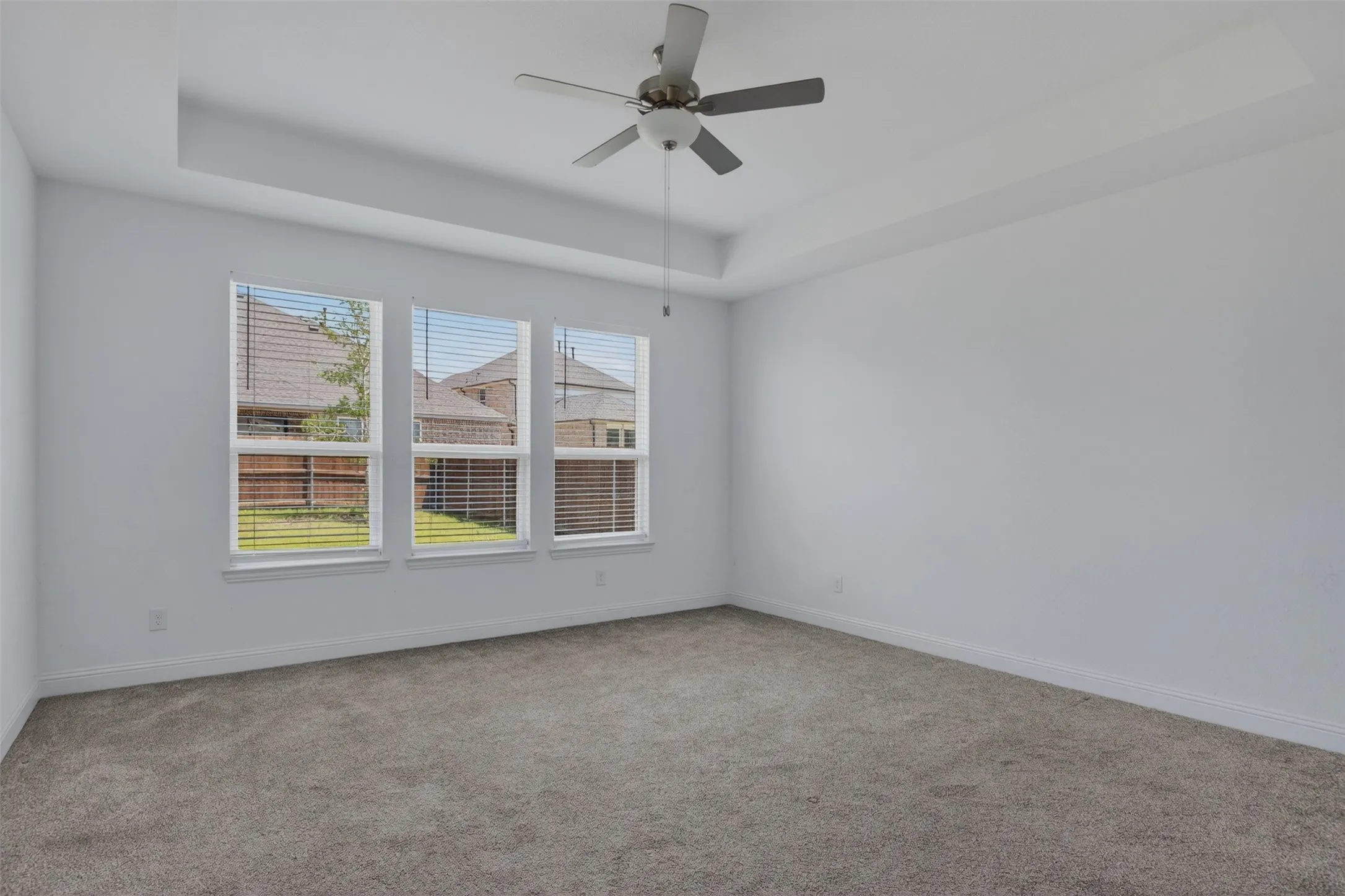 Spacious room featuring a tray ceiling, ceiling fan, three large windows with blinds, plush gray carpeting, and white baseboards