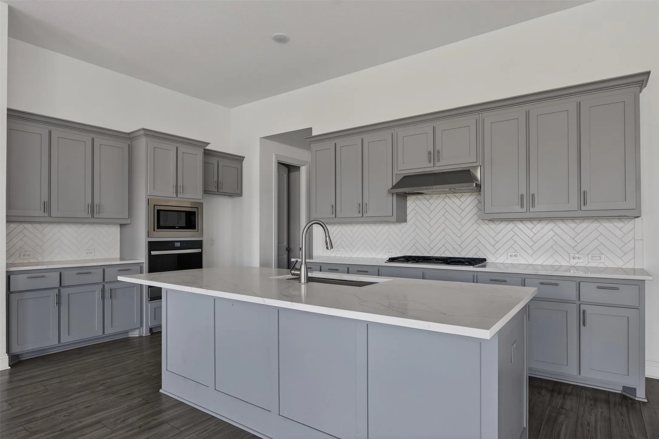 Kitchen featuring gray shaker-style cabinetry, a central island with an undermount sink, stainless steel appliances, a herringbone tile backsplash, and wood-finish flooring