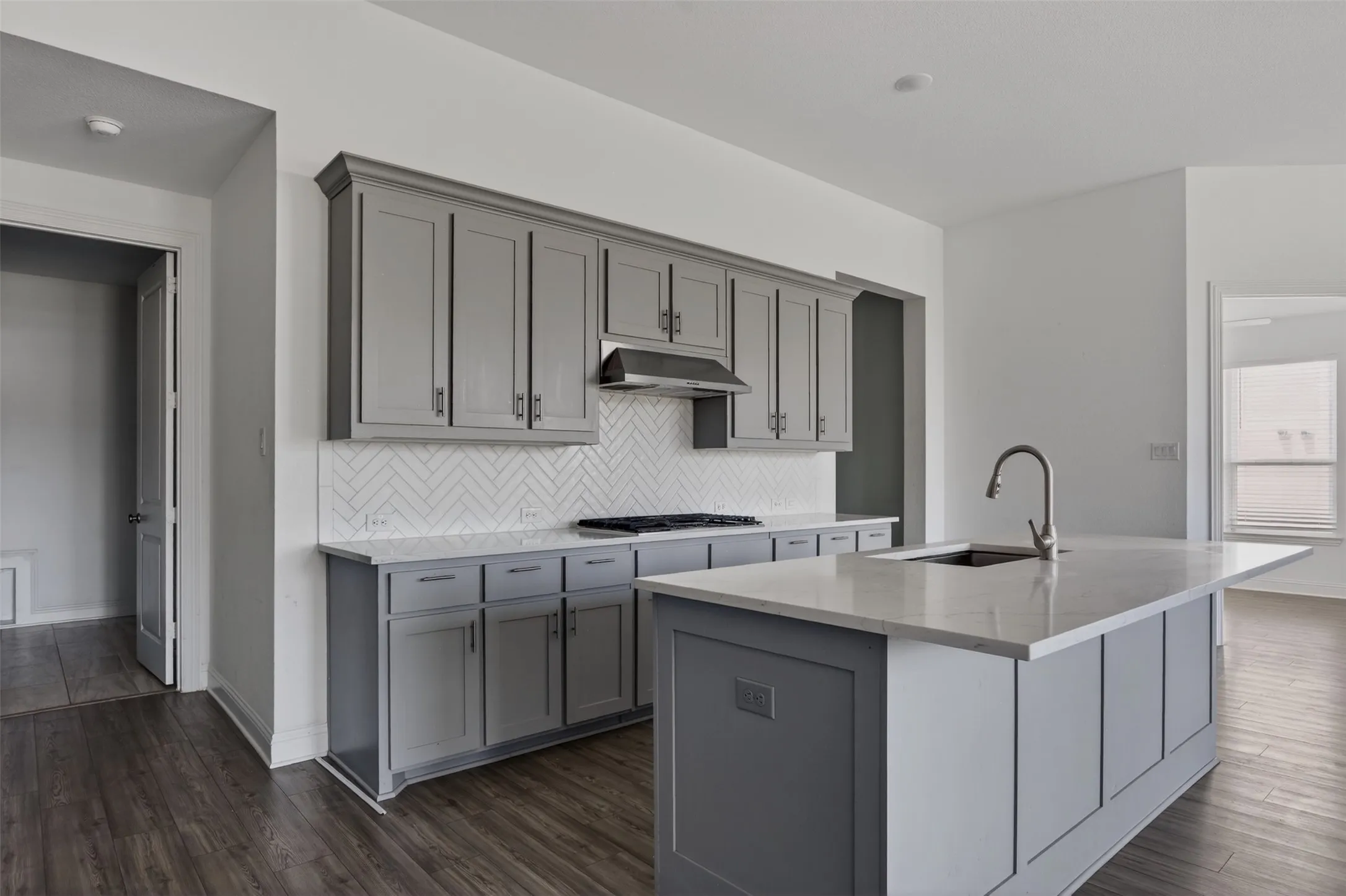 Modern kitchen featuring grey shaker cabinetry, white herringbone tile backsplash, a center island with a built-in sink and gooseneck faucet, light-toned countertops, and wood-finish flooring