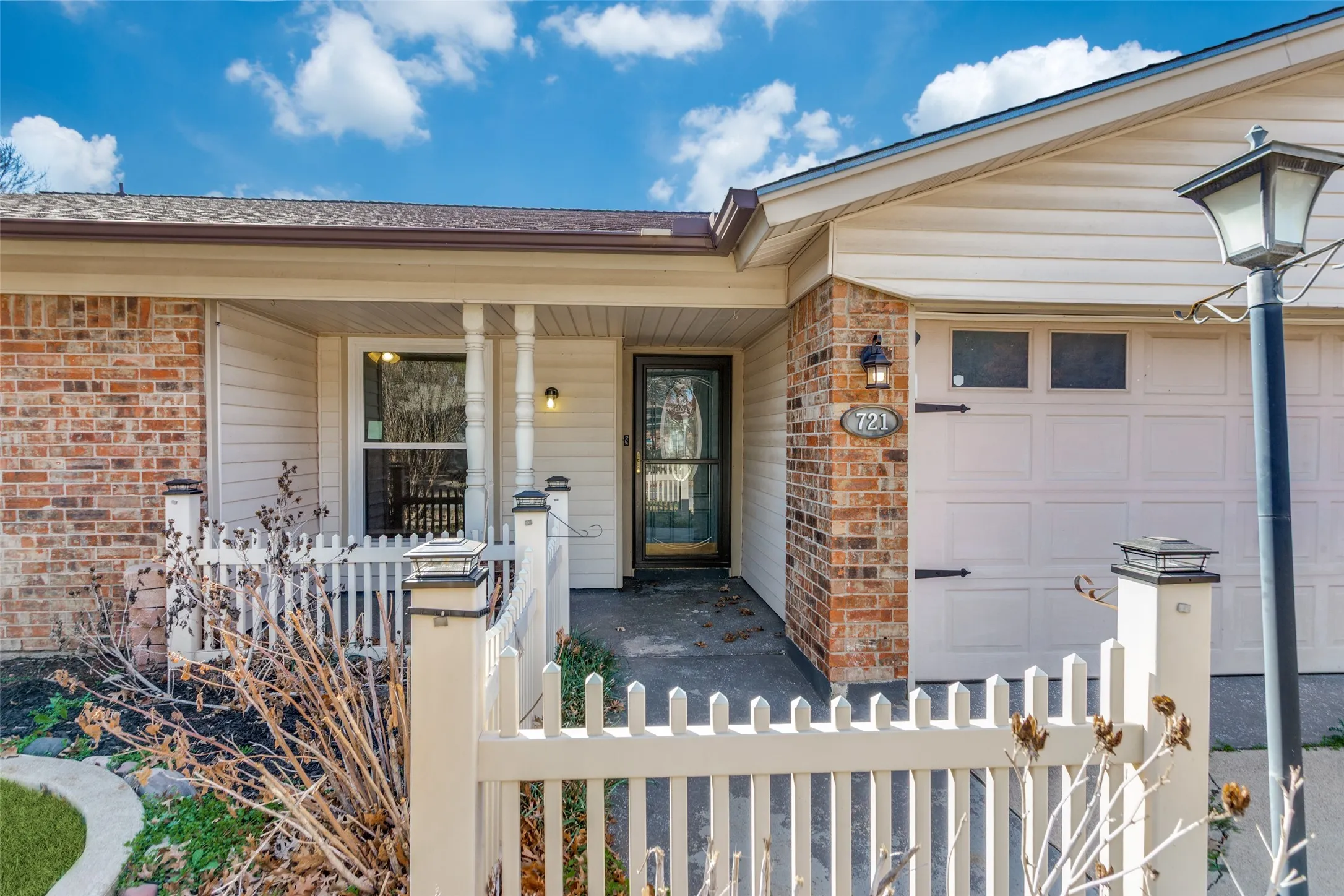 Brick and siding exterior featuring a covered porch with white columns, an attached garage with decorative hardware, a white picket fence, and exterior lighting fixtures