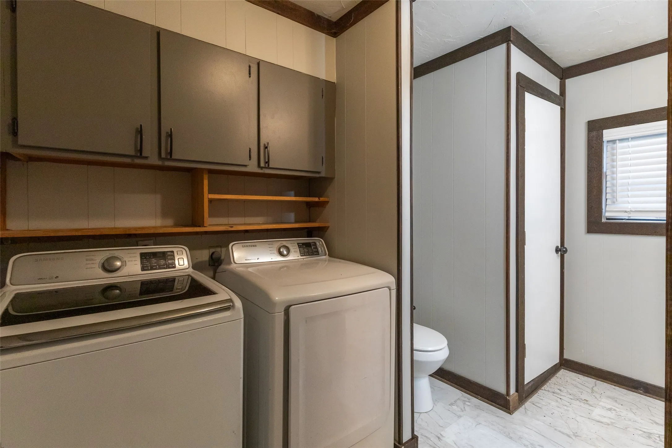 Utility area featuring upper cabinetry, open shelving, a white paneled wall, a privacy partition, and window with blinds