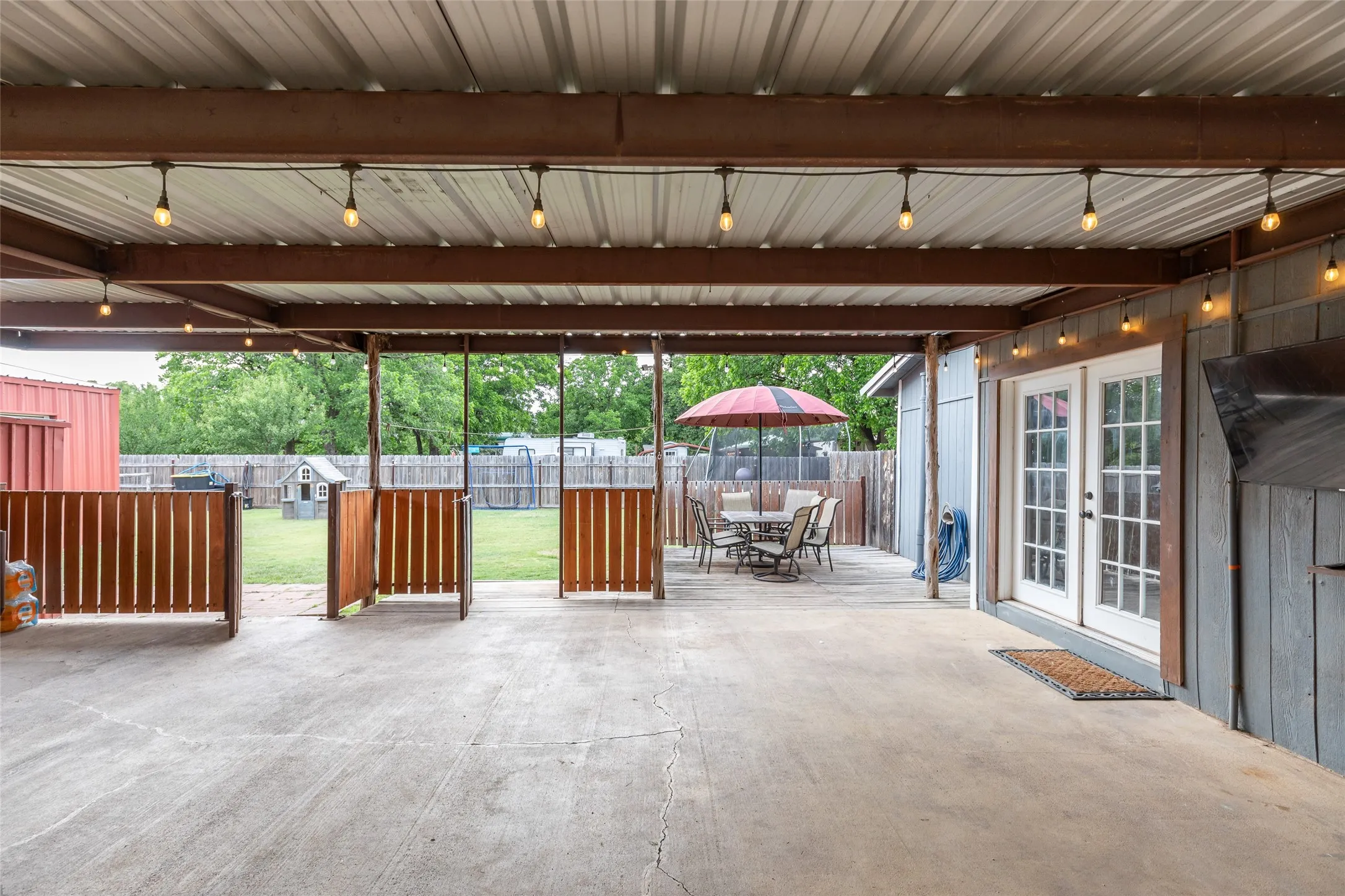 Covered concrete patio featuring string lighting, wood fencing, and a metal roof structure
