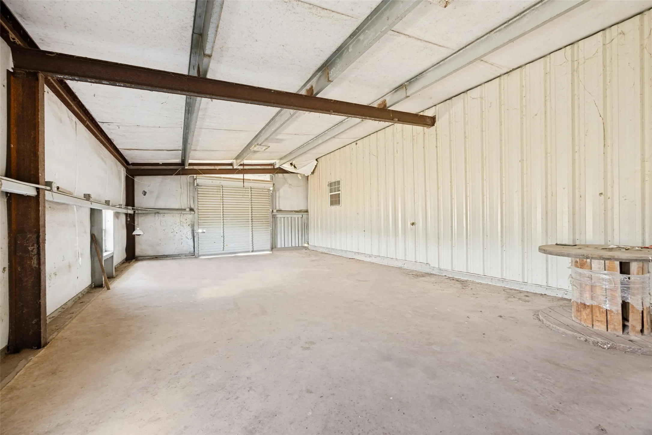 Spacious interior featuring a concrete floor, exposed structural beams, corrugated metal walls, a roll-up door, and an operable window