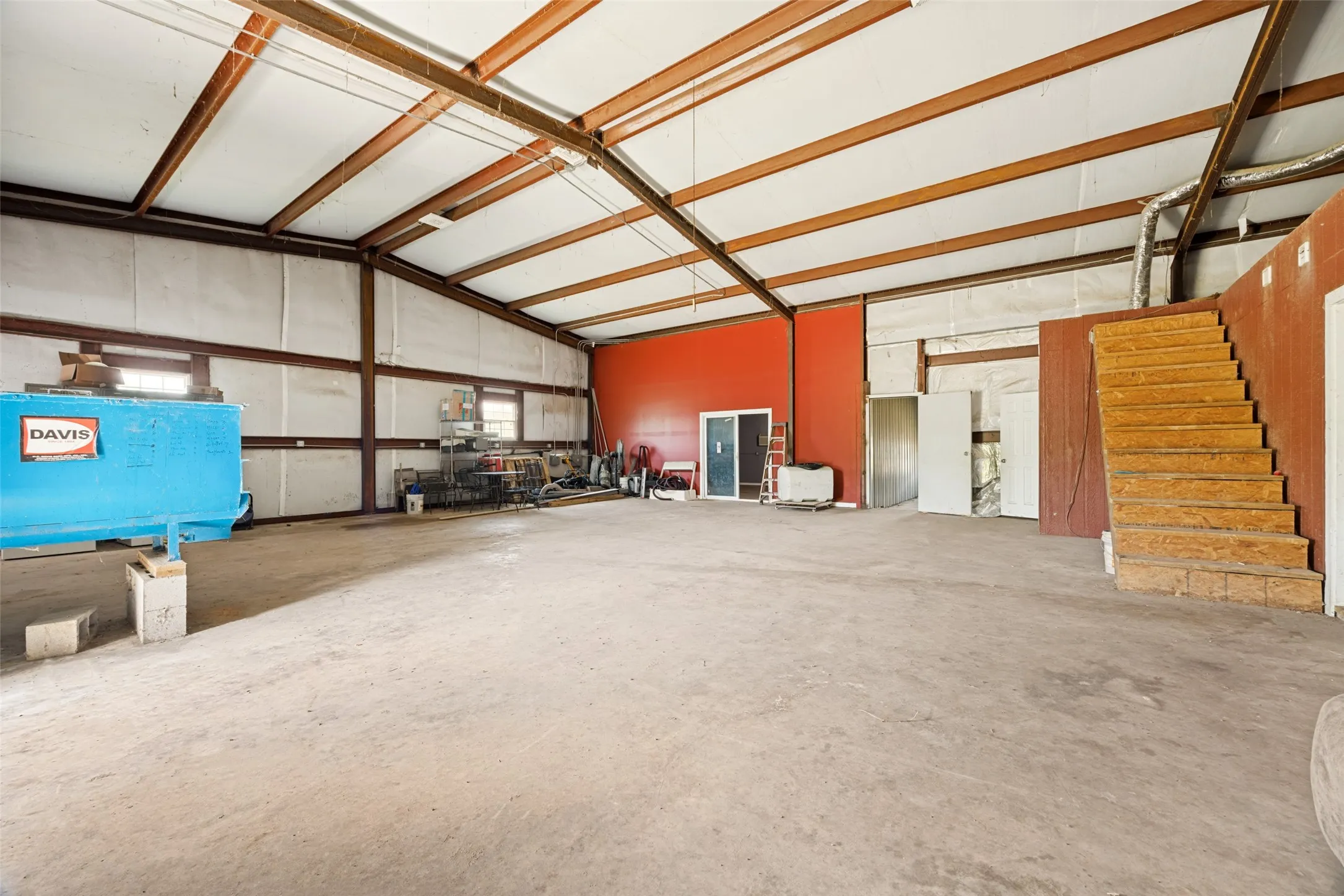 Spacious industrial interior featuring a concrete floor, exposed metal beam ceiling structure, and a partial second-story loft accessed by a wood staircase
