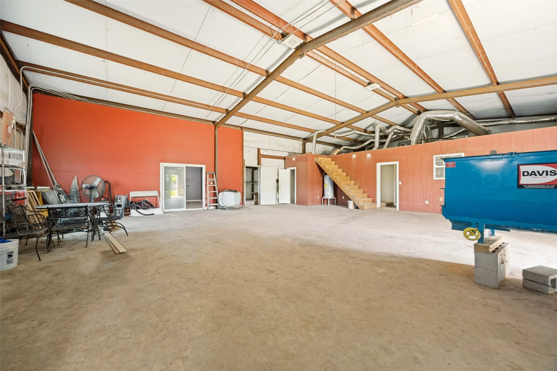 Spacious open-plan metal building featuring a concrete floor, exposed structural steel beams, high ceilings, and an internal upper-level structure with a built-in staircase