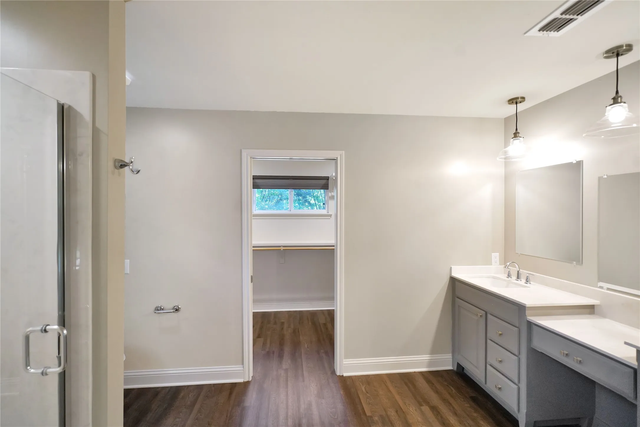 Spacious bathroom featuring wood-finish flooring, a dual-sink vanity with light gray cabinetry, white countertops, and contemporary pendant lighting