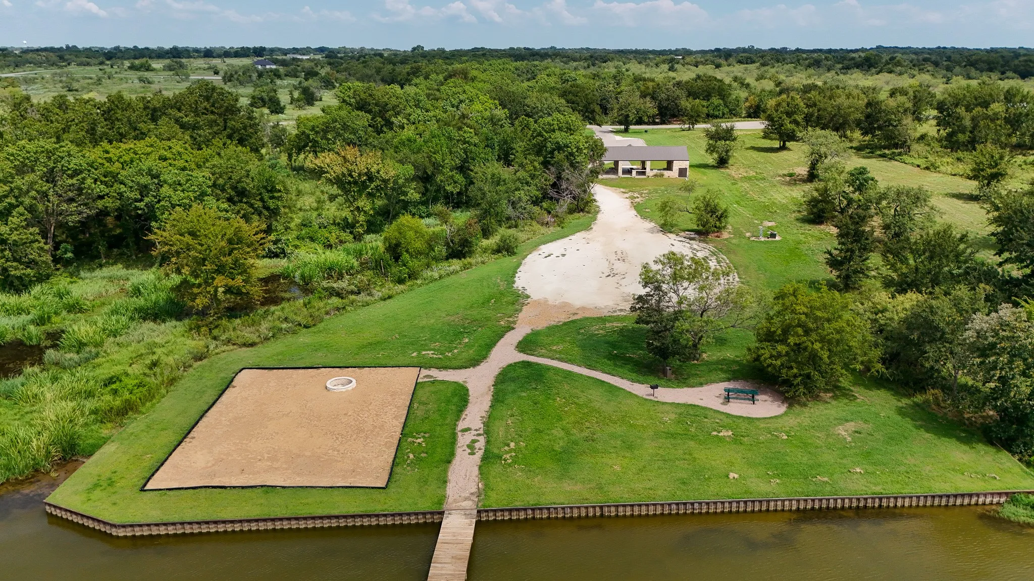 Expansive green space featuring a sand court with a central fire pit, a wooden pier, and a winding path leading to a covered pavilion
