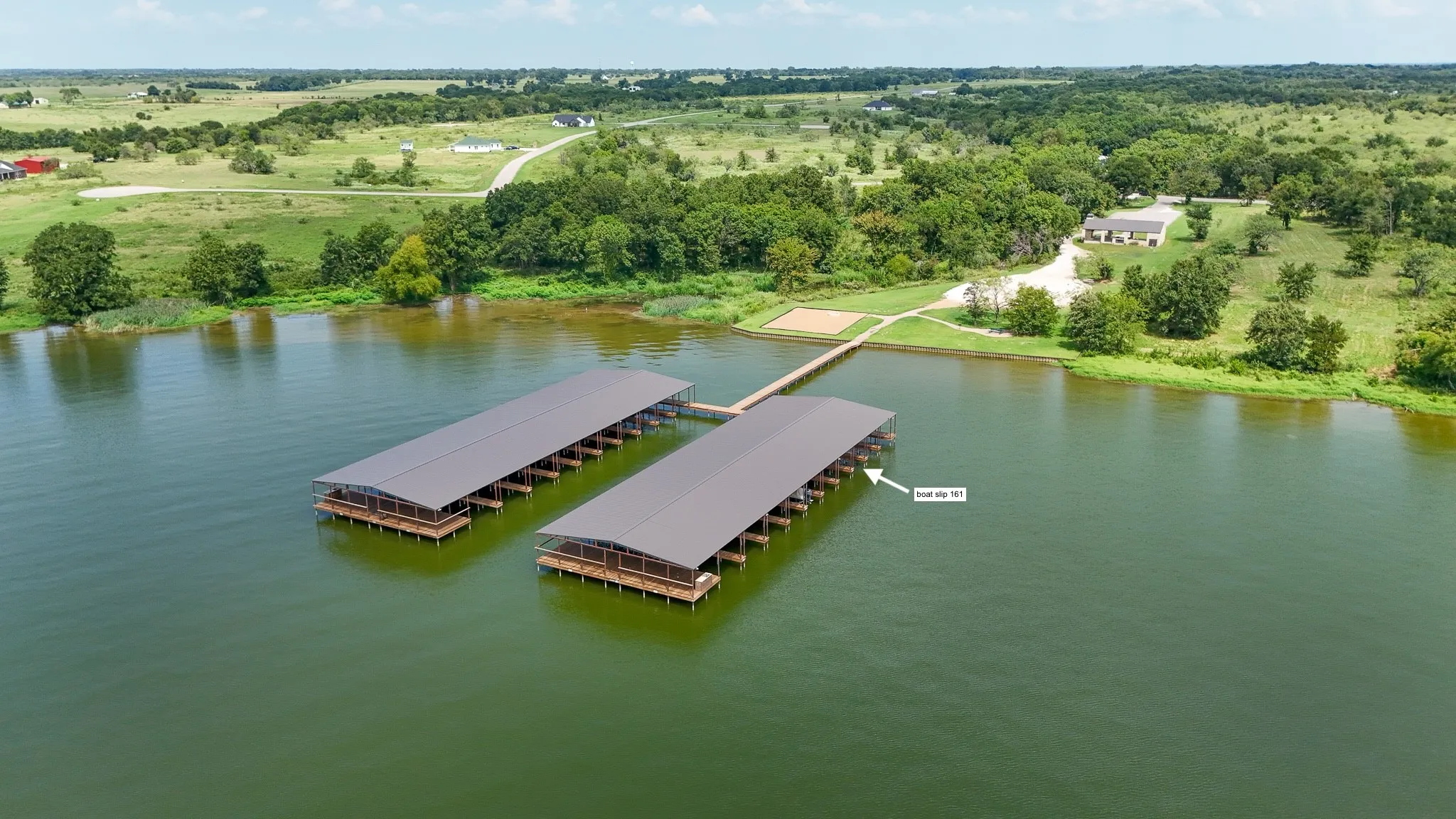 Two covered boat docks extending into the lake, featuring multiple slips, a connecting walkway, and surrounding green landscapes