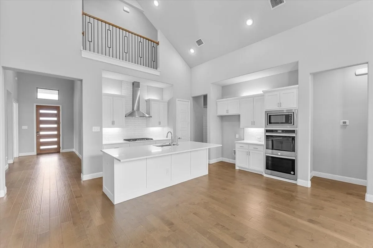 Open-concept kitchen featuring an island with sink, white shaker cabinetry, stainless steel appliances, and wood-finish flooring