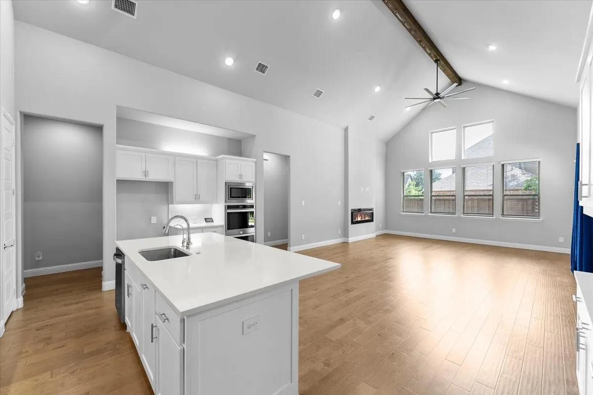 Open-concept living area featuring a kitchen island with sink, integrated double ovens, wood-finish flooring, a contemporary fireplace, and vaulted ceilings with an exposed beam