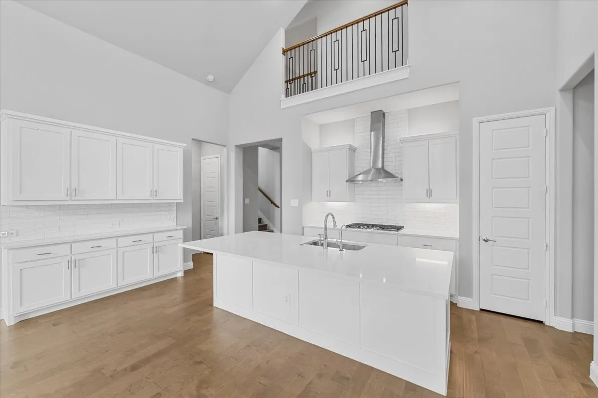 Spacious kitchen featuring an expansive central island with an integrated sink, white shaker cabinetry, subway tile backsplash, stainless steel range hood, and wood-finish flooring