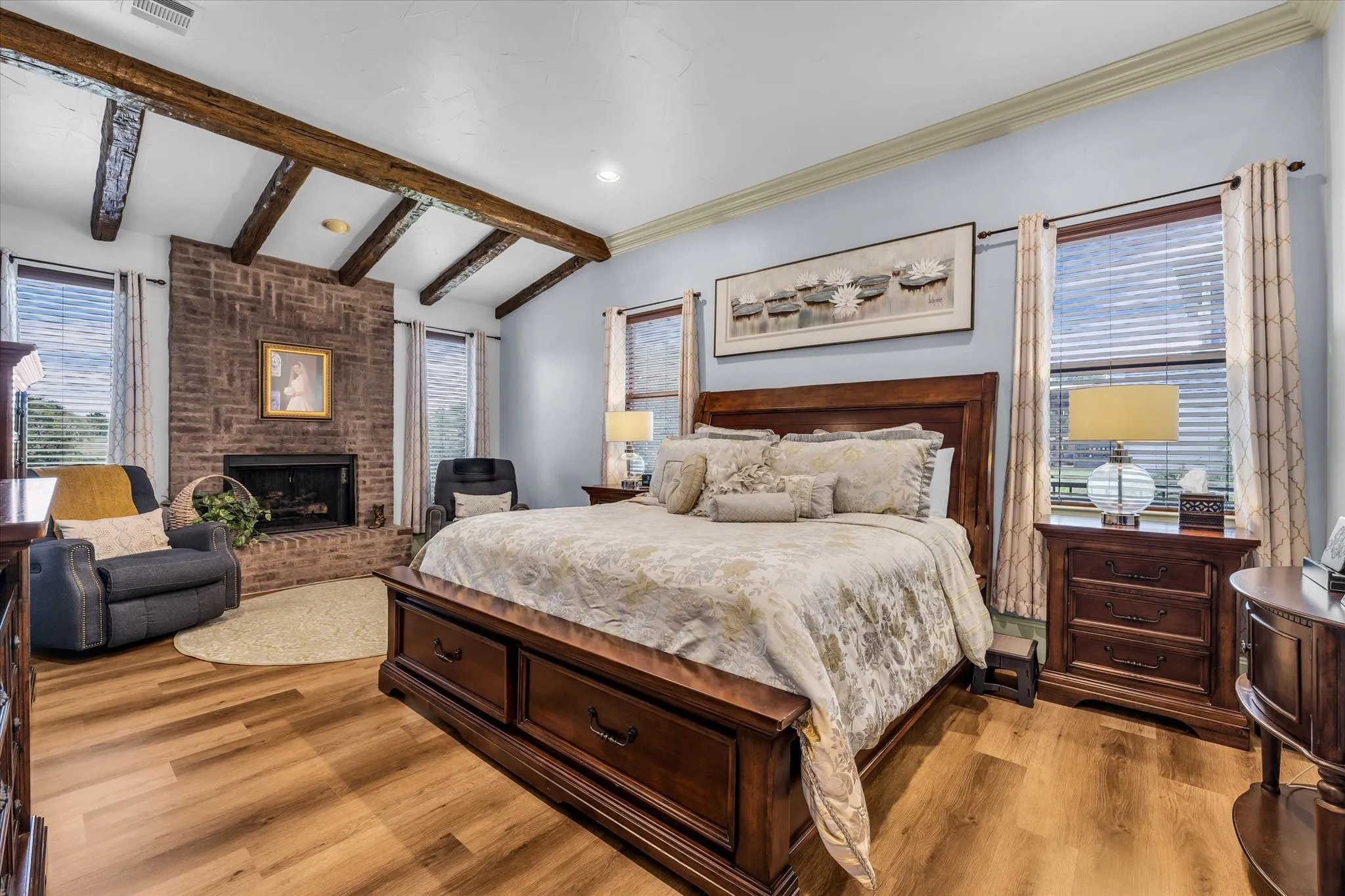 Spacious room featuring vaulted ceilings with exposed wood beams, a brick fireplace, wood-finish flooring, and multiple windows with blinds