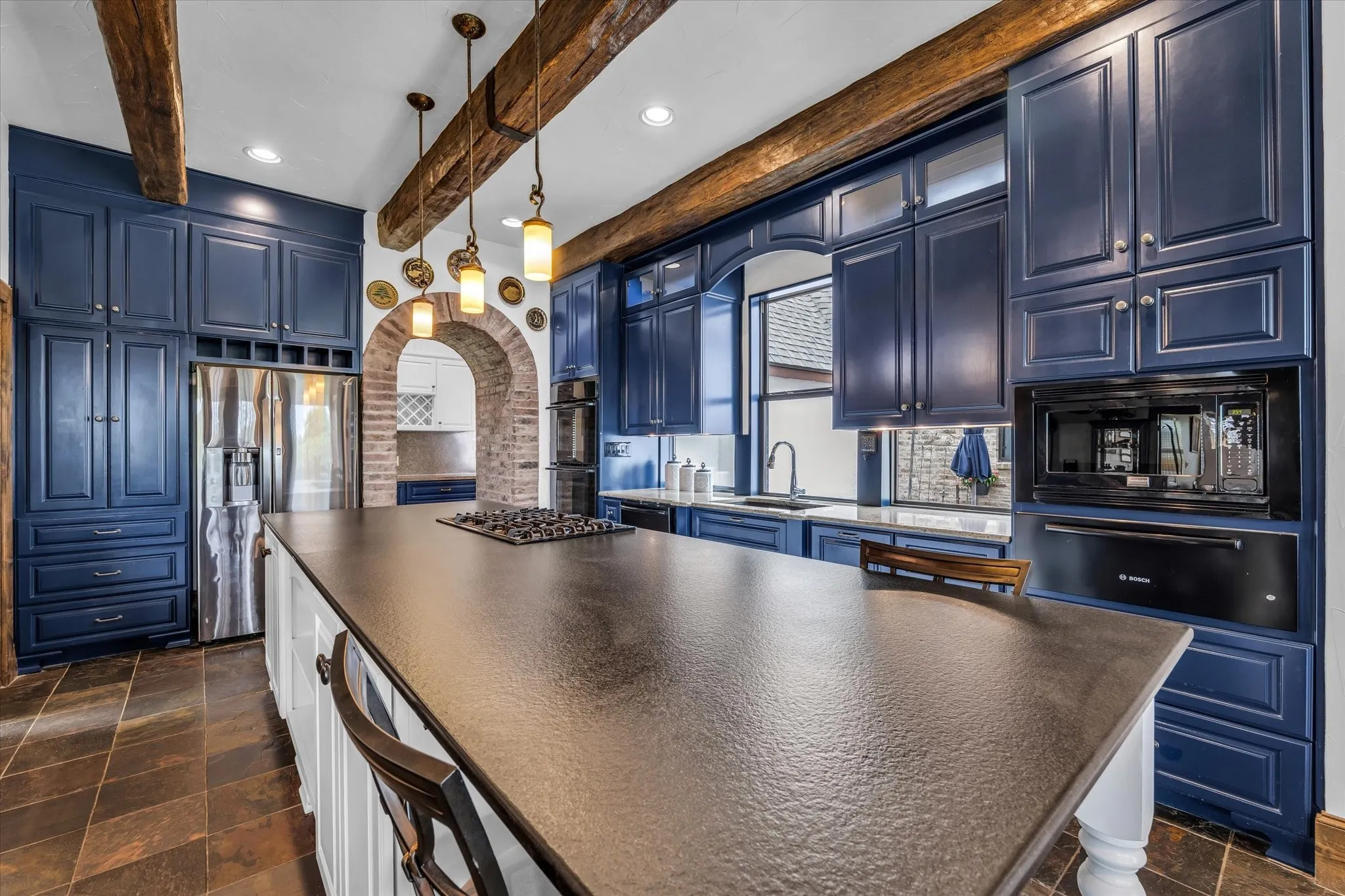Spacious kitchen featuring an expansive center island with an integrated cooktop, blue cabinetry, slate tile flooring, and rustic exposed beams