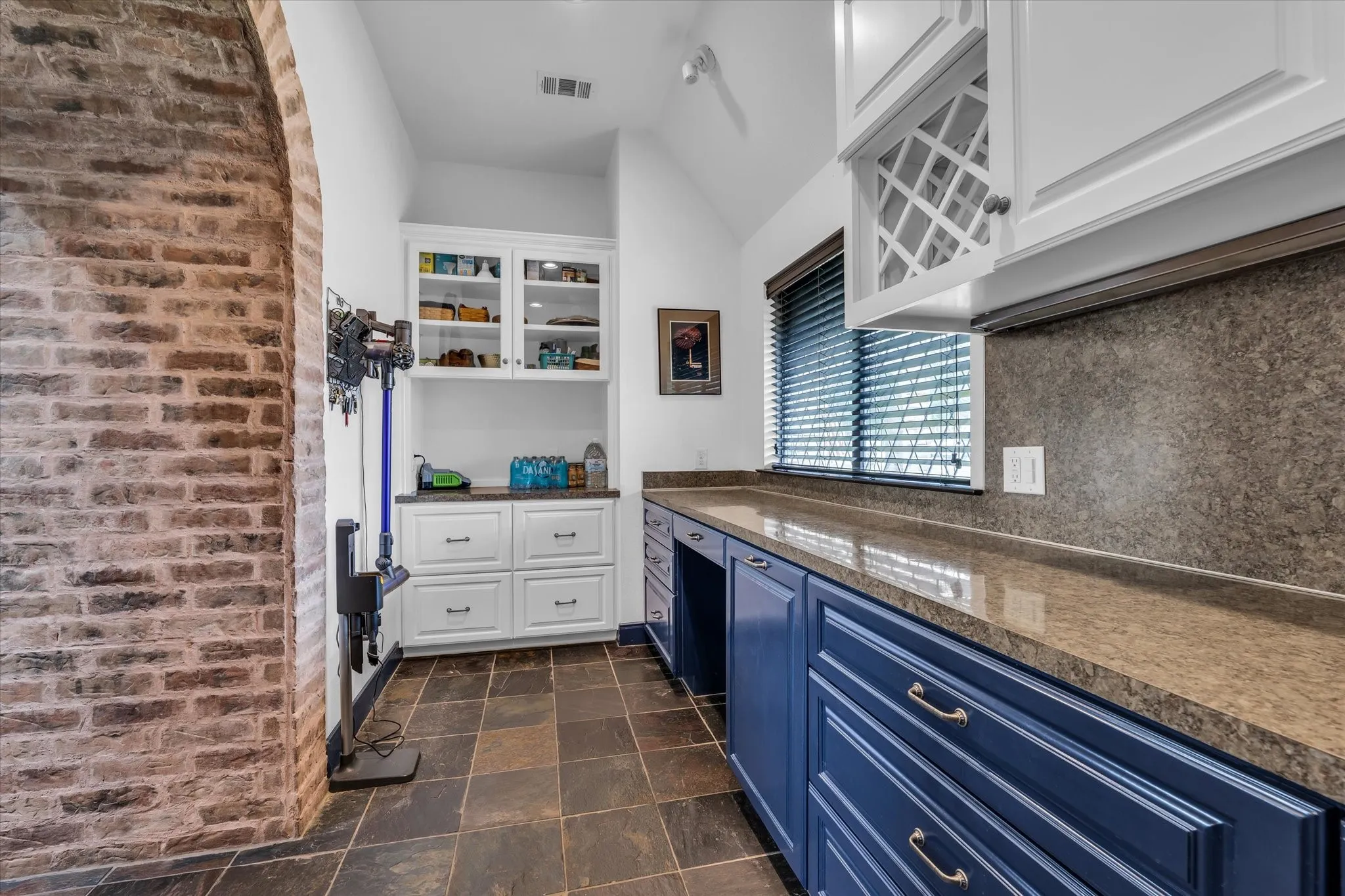 Utility space featuring a brick archway, slate tile flooring, extensive countertop surfaces, built-in cabinetry with glass-front uppers, and a window with blinds