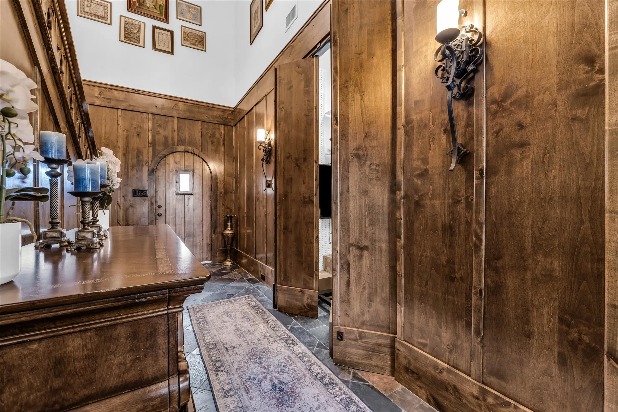 Hardwood paneling in a warm finish, arched entry door with a small window, dark slate tile flooring, wall-mounted decorative sconces, and a finished wood staircase with a matching railing