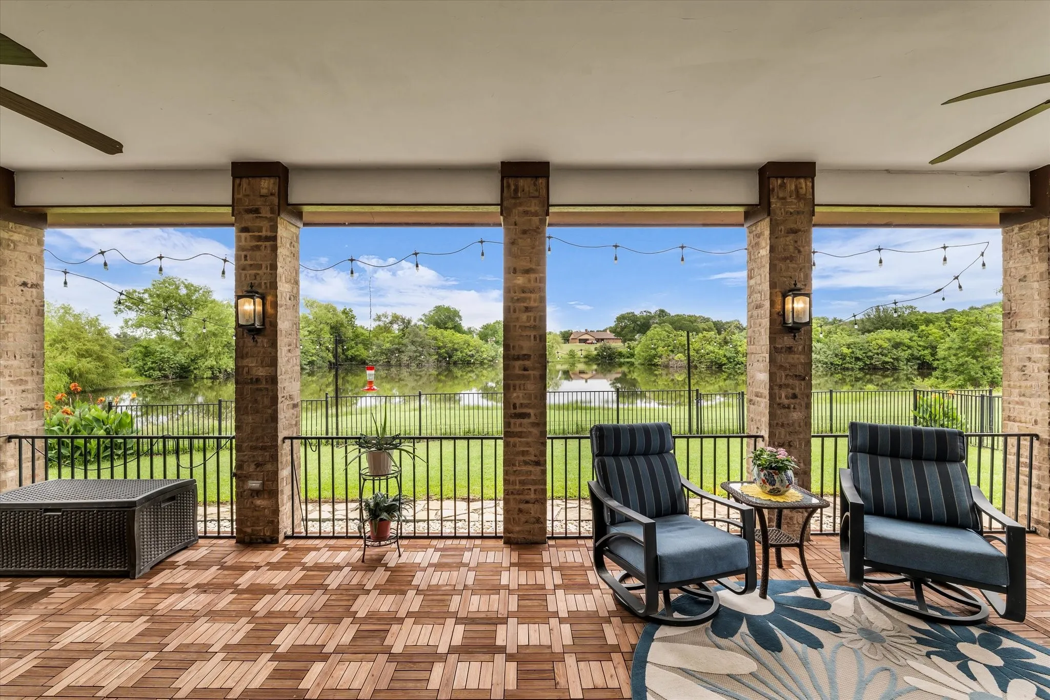 Covered patio with wood-finish flooring, brick columns, and a black wrought iron railing overlooking a body of water