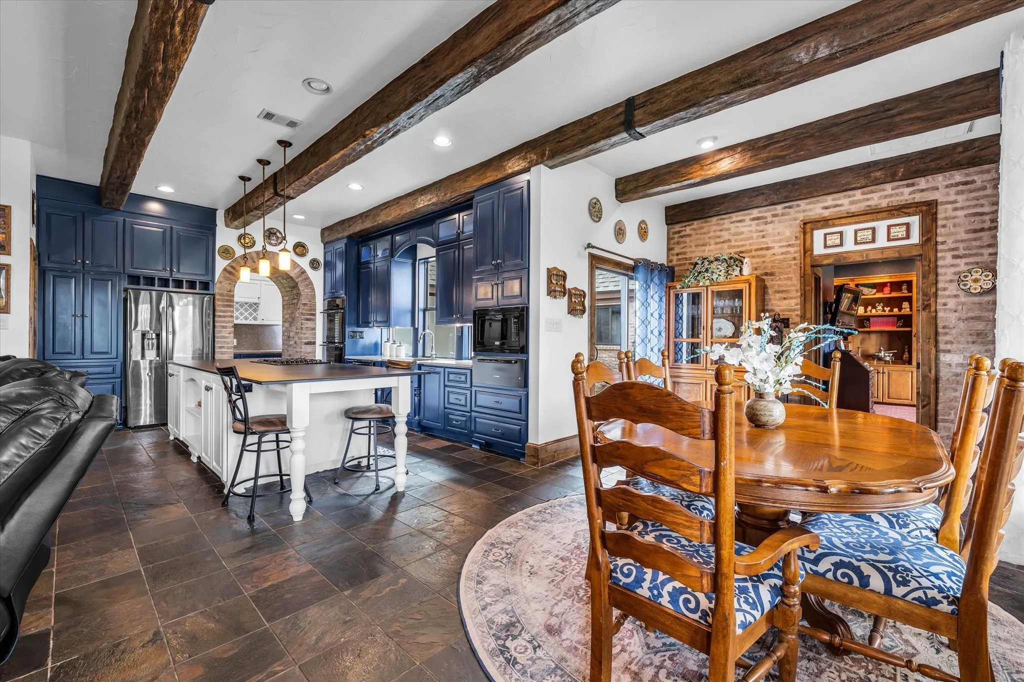 Spacious kitchen and dining area featuring exposed wood beams, slate tile flooring, blue cabinetry, a central kitchen island with seating, and a brick accent wall