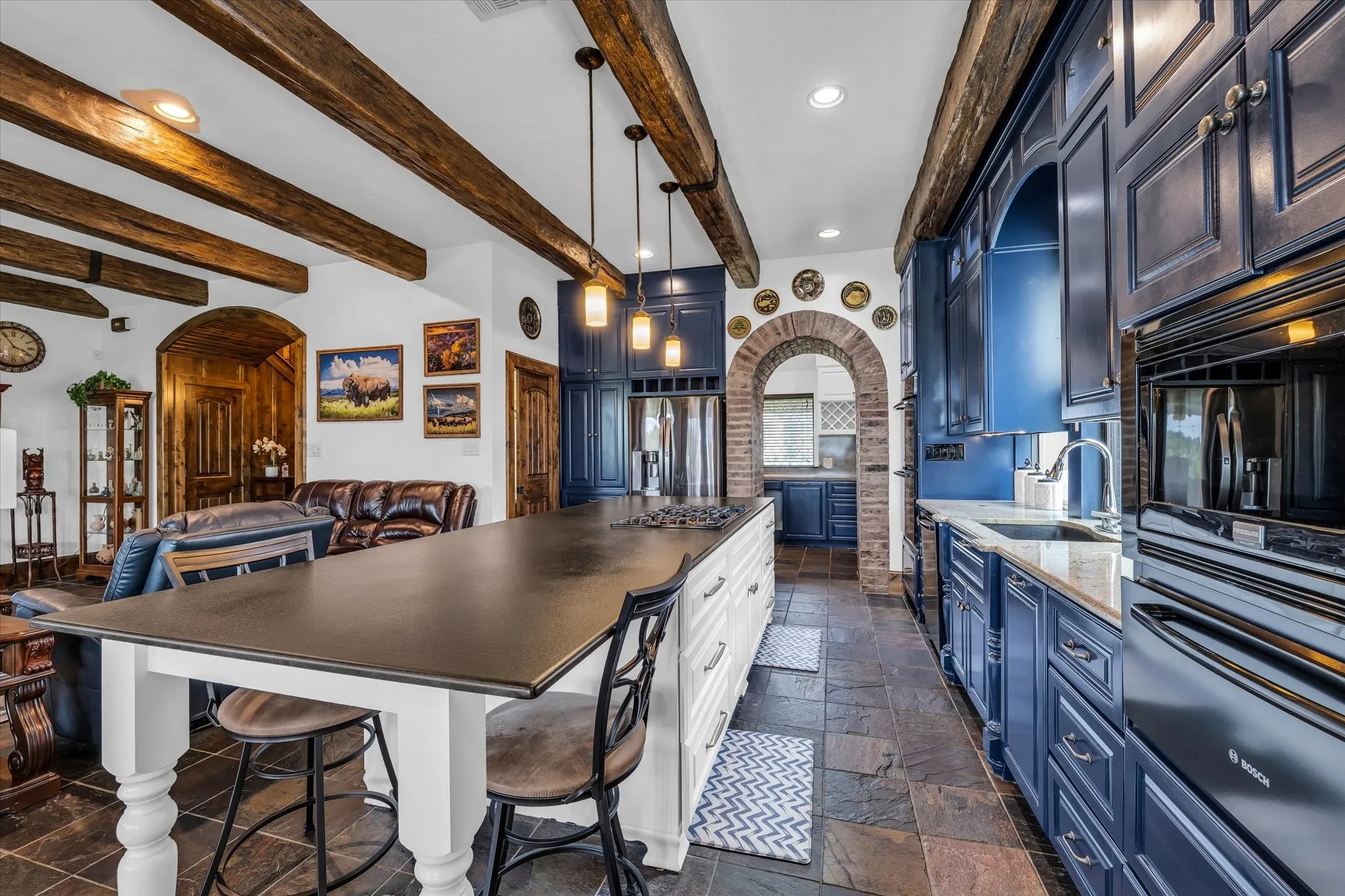 Spacious kitchen featuring deep blue cabinetry, stone tile flooring, a large center island with seating, built-in appliances, and a brick arched doorway
