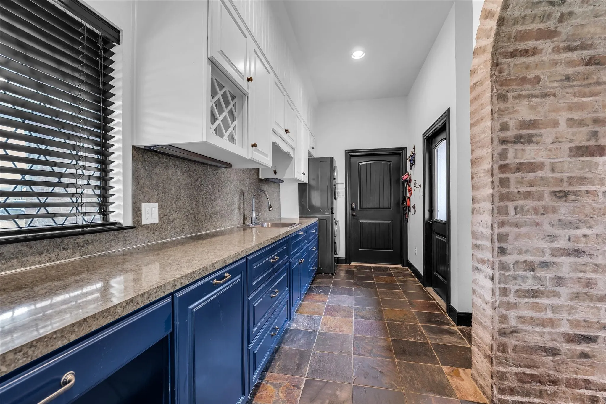 Utility space featuring slate-style floor tile, white upper cabinetry, blue lower cabinetry, stone countertops, and a full-height stone-look backsplash