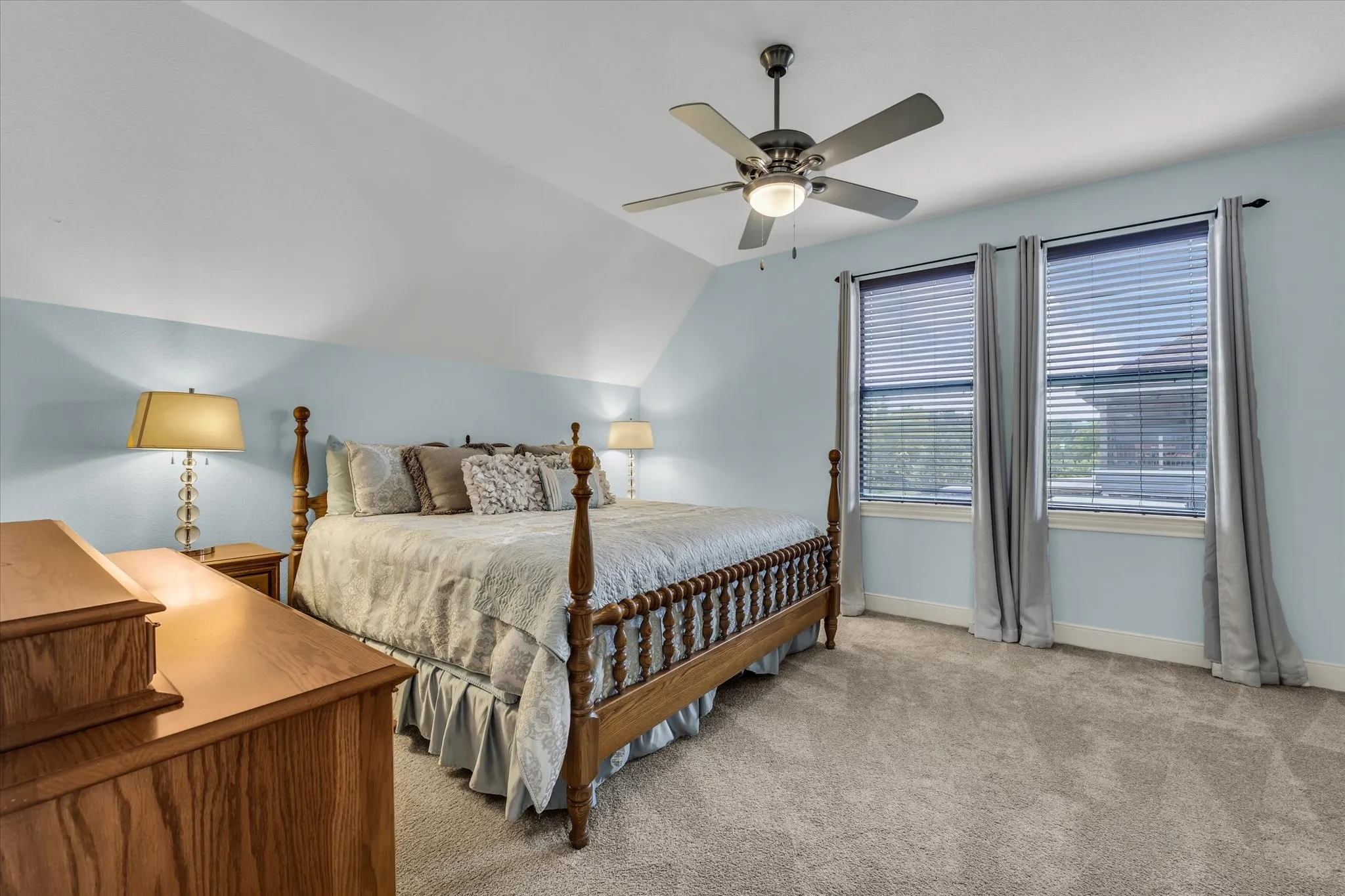 Bedroom featuring vaulted ceilings, light blue walls, two windows with blinds, and carpet flooring