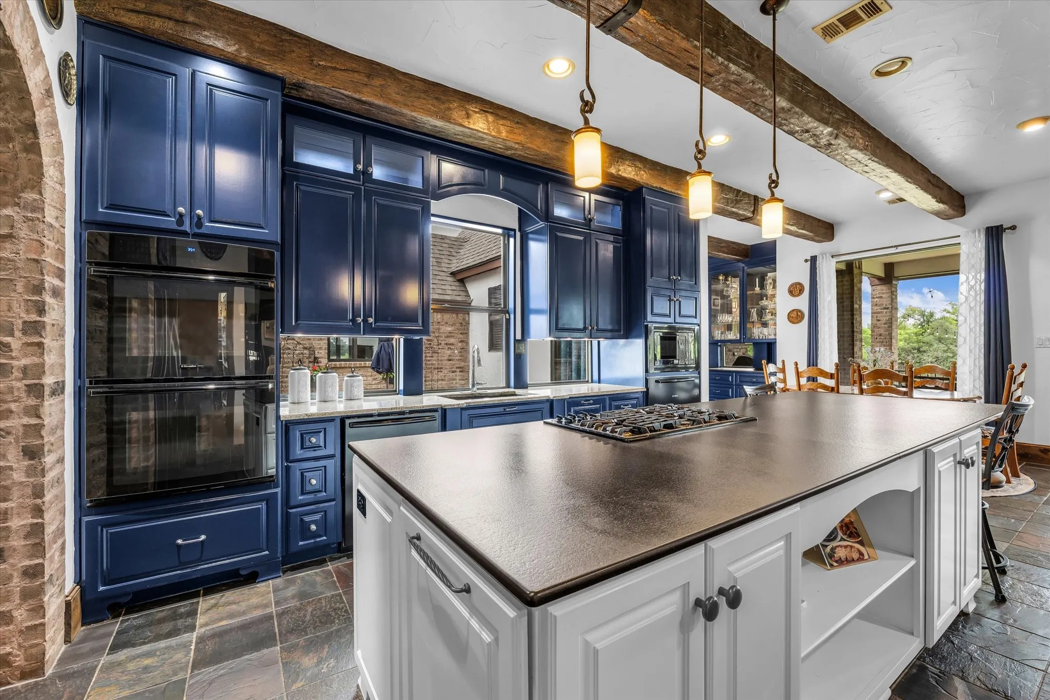 Kitchen featuring an oversized central island with a built-in cooktop, rich blue cabinetry, double wall ovens, exposed timber beams, and natural stone flooring