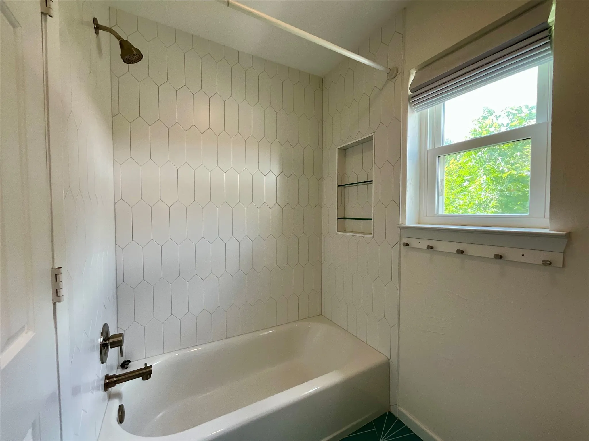 Bathtub and shower with white picket tile surround, bronze fixtures, built-in shelving, and a window with a fabric shade