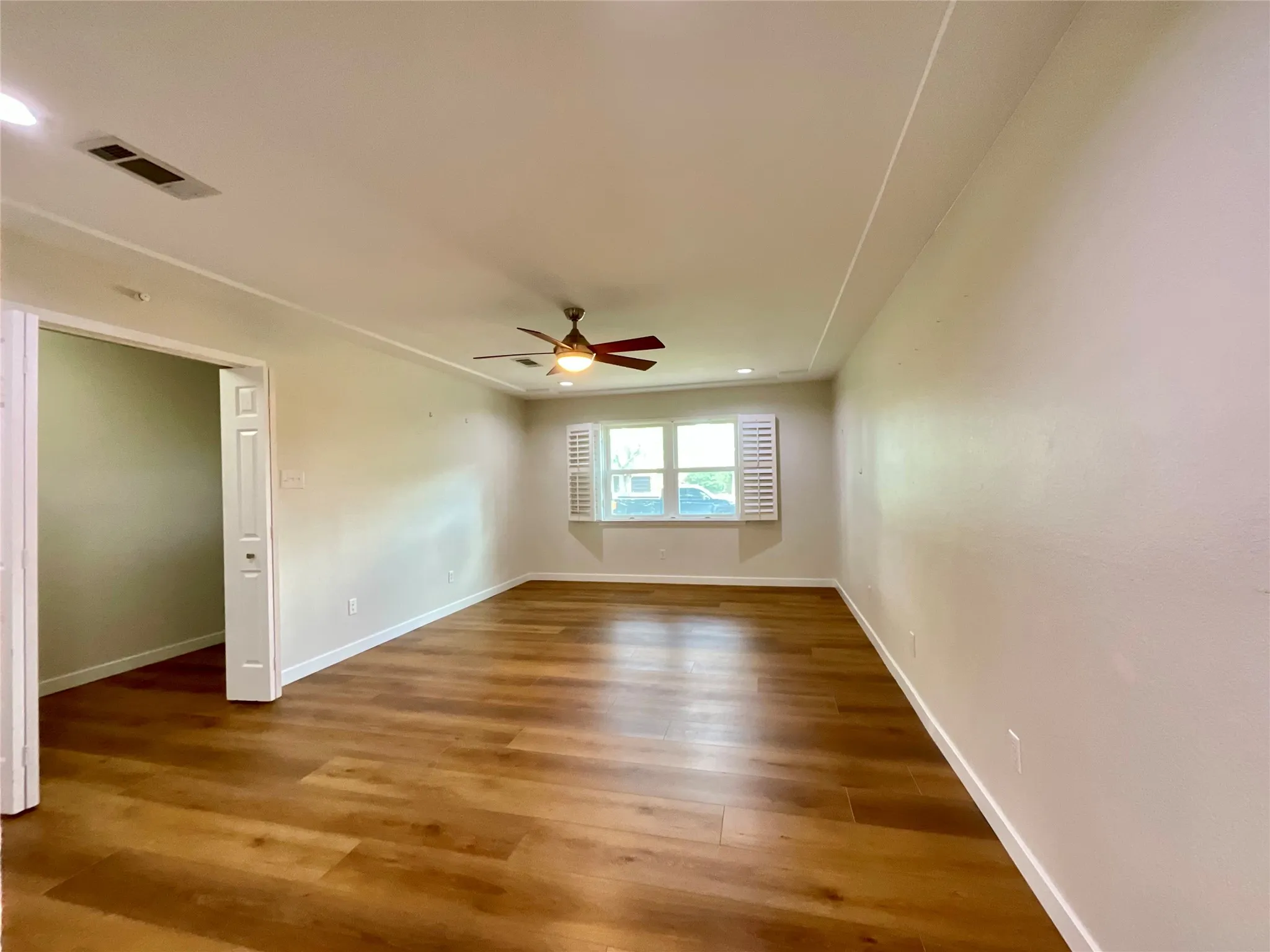 Expansive room featuring wood-finish flooring, a ceiling fan with integrated lighting, recessed lighting, and a large window with plantation shutters