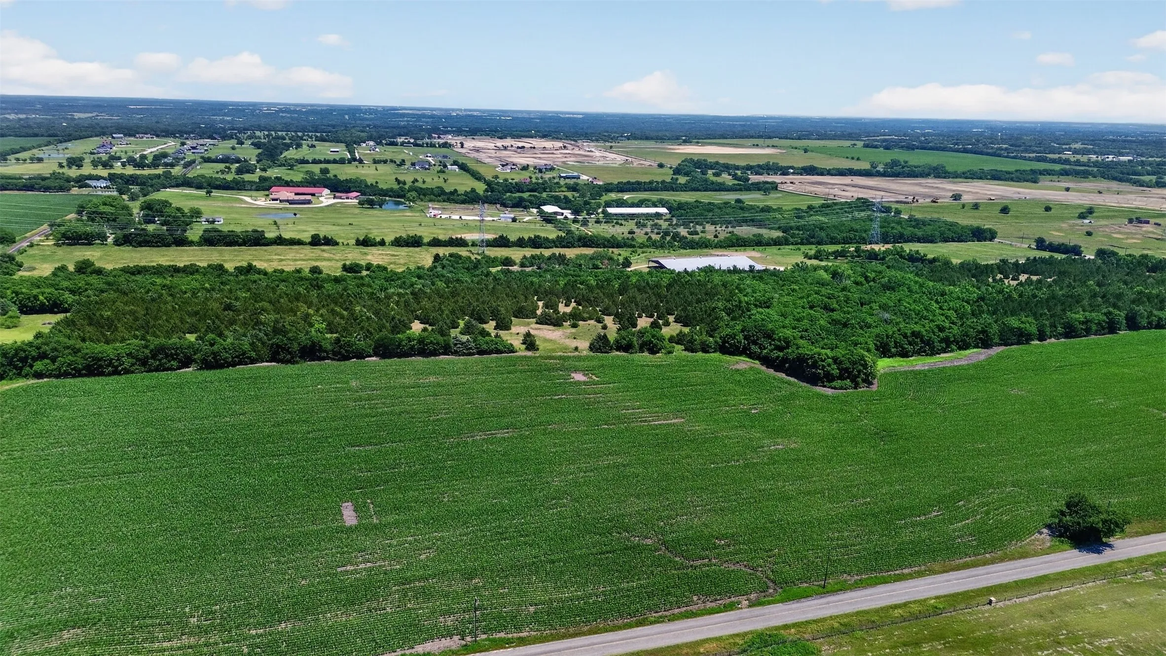 Expansive green fields bordering a paved road, featuring scattered wooded areas and distant structures under a clear sky