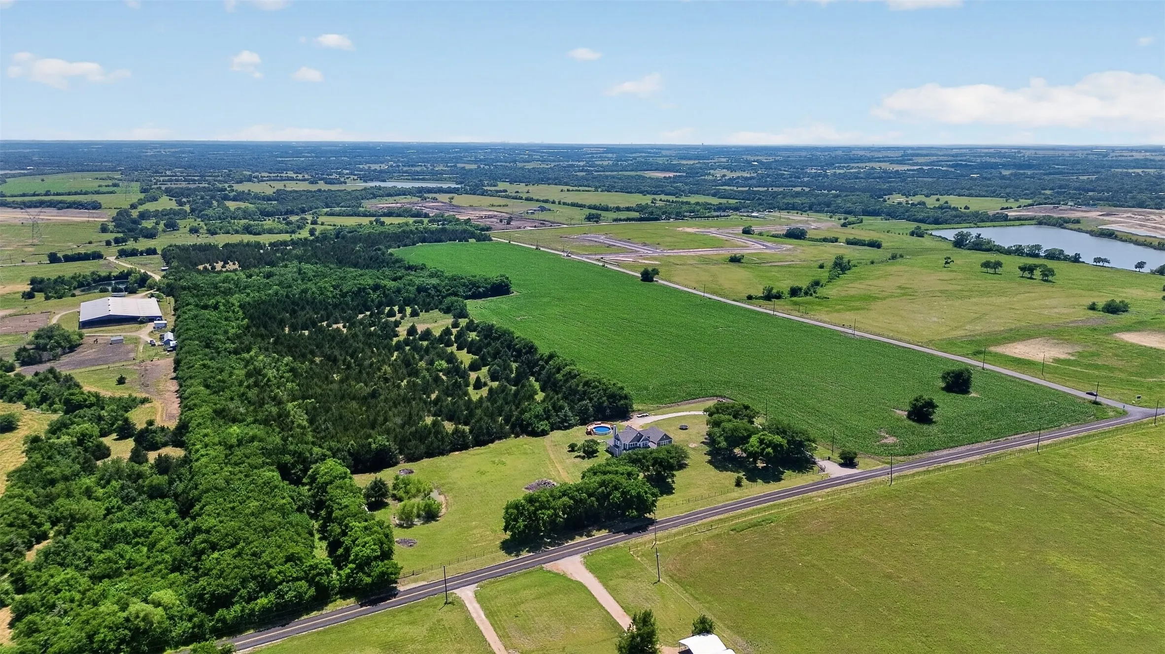 Expansive rural property featuring a main residence with a pool, surrounded by mature trees, open green fields, and a nearby body of water