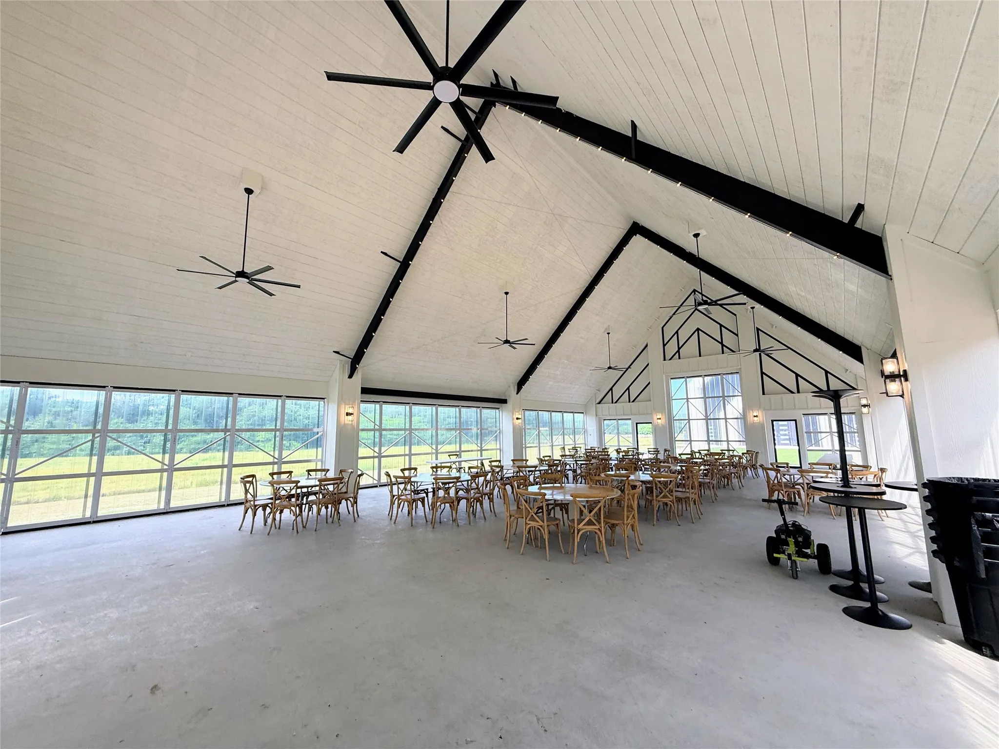 Spacious interior featuring a vaulted ceiling with exposed black beams, white shiplap ceiling panels, large industrial-style ceiling fans, and an expansive wall of glass garage doors
