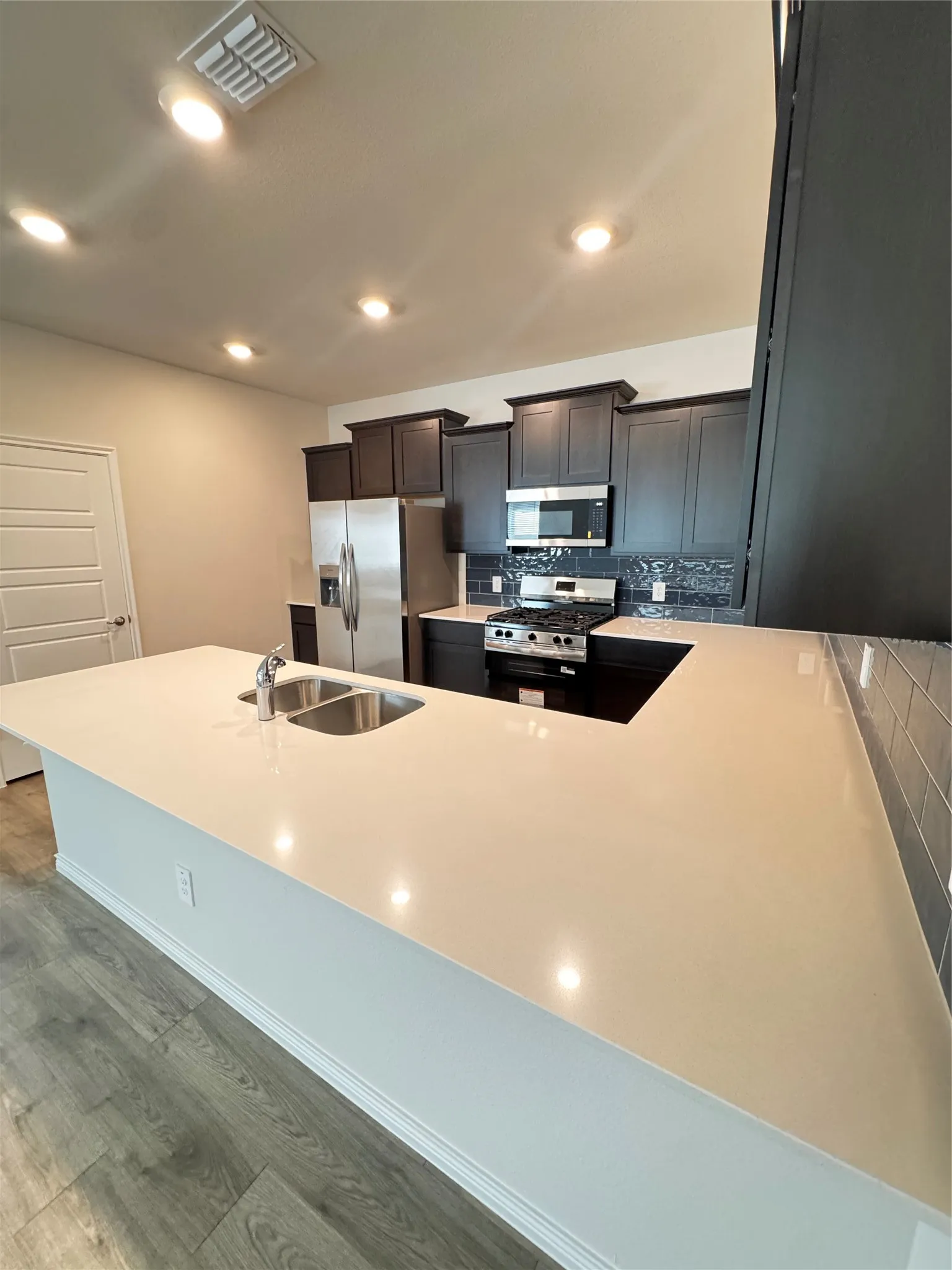 Kitchen featuring a spacious island with an integrated double sink, stainless steel appliances, dark wood cabinetry, subway tile backsplash, and recessed lighting