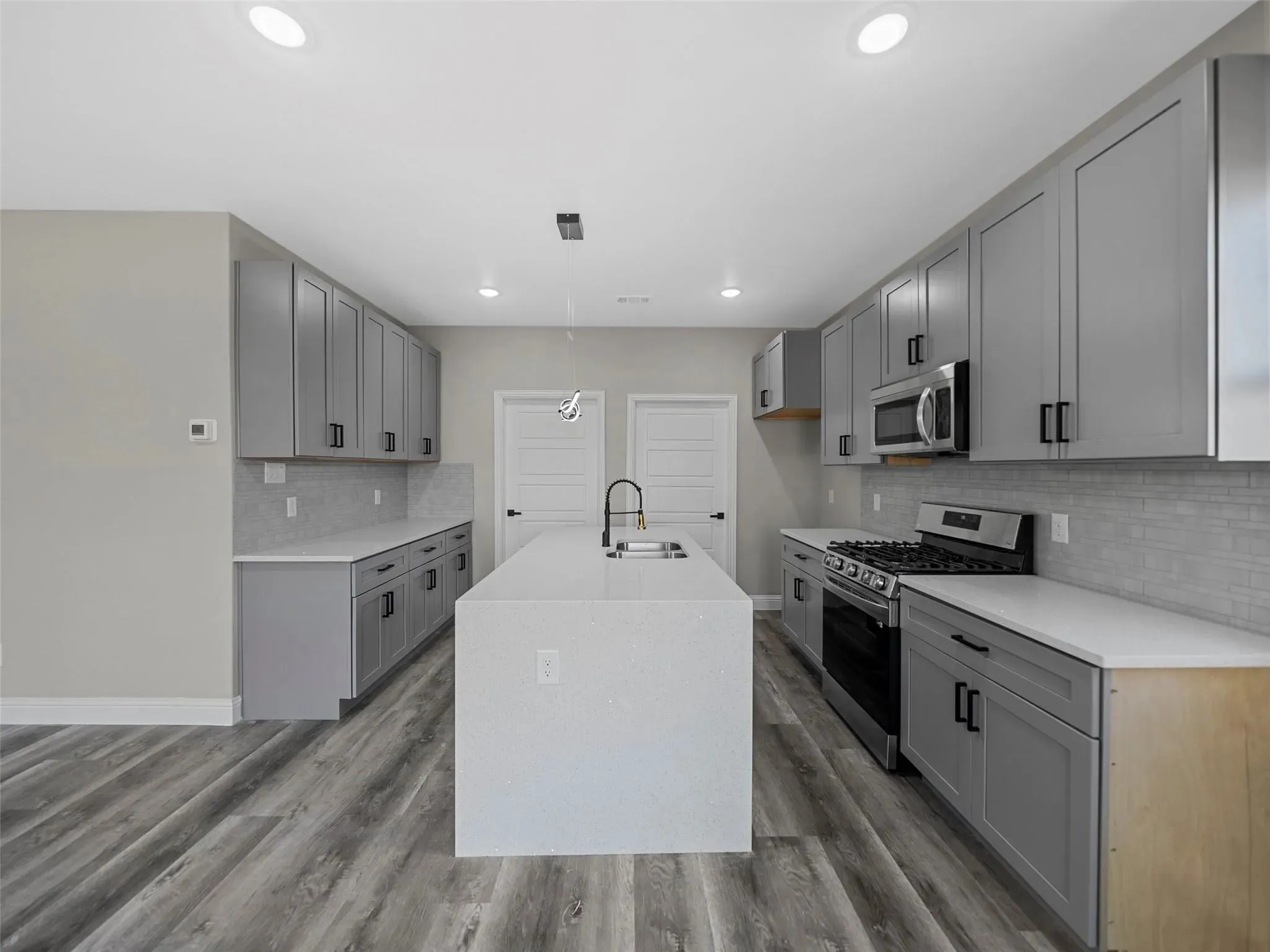Modern kitchen featuring light gray cabinetry, a central island with an integrated sink, stainless steel appliances, white countertops, and wood-finish flooring