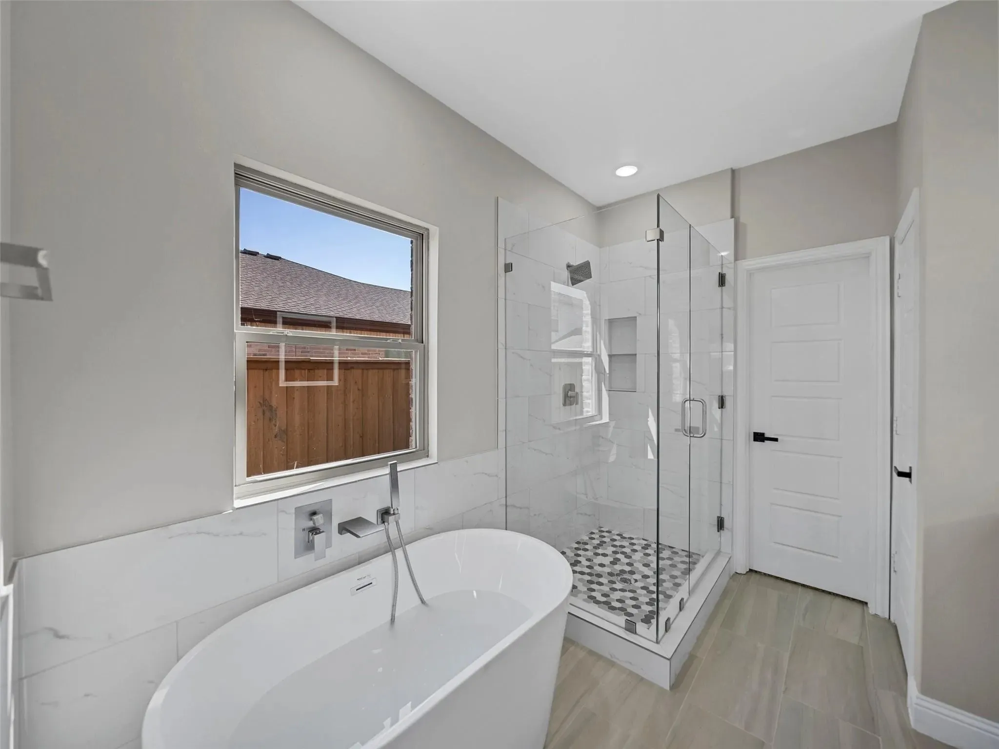 Soaking tub with wall-mounted chrome faucet, glass-enclosed shower with tiled walls and pebble flooring, and light wood-finish flooring