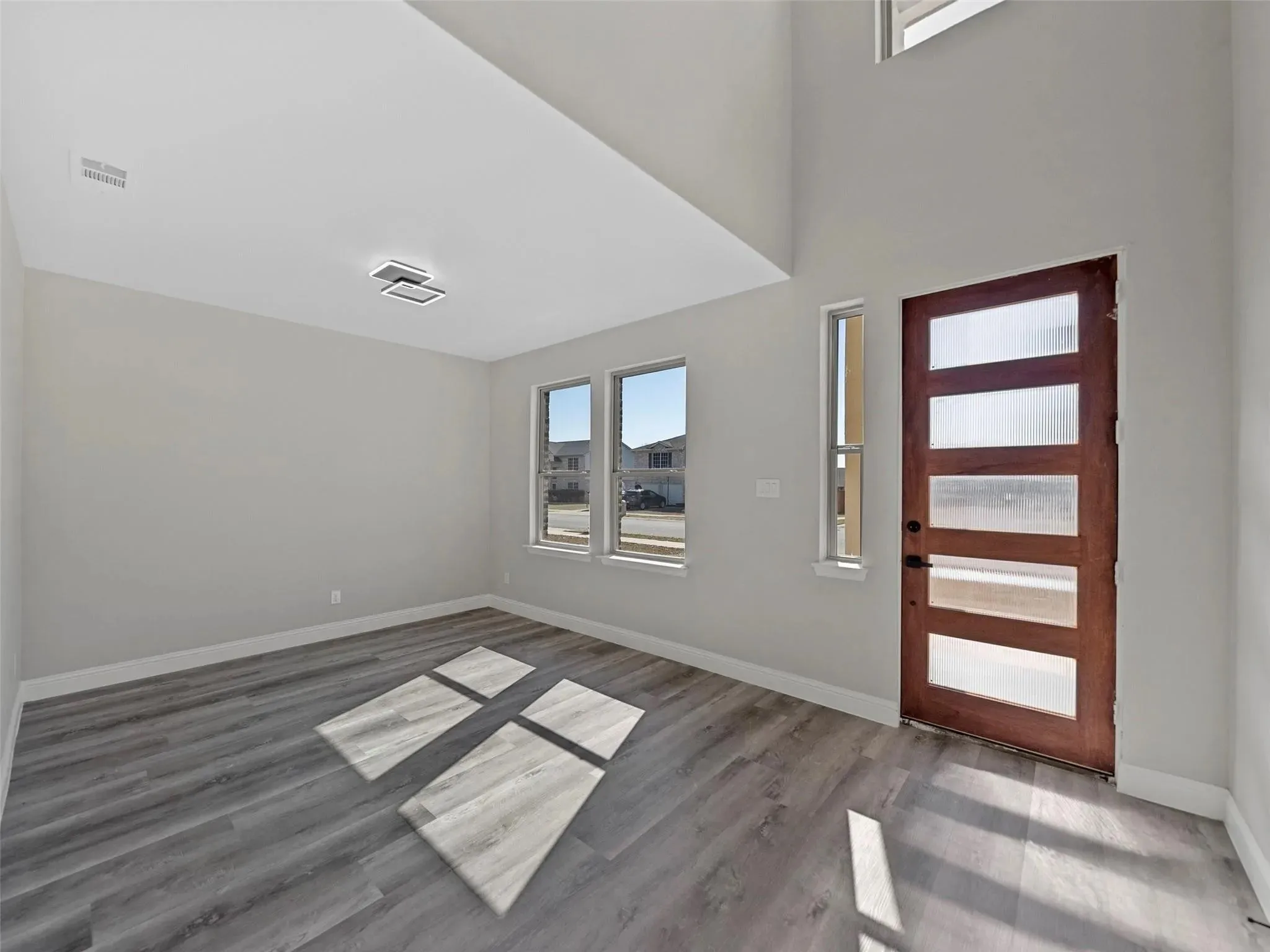 Entry foyer featuring a modern wood-finish door with frosted glass panels, gray wood-finish flooring, high ceilings, multiple windows, and a contemporary flush-mount light fixture