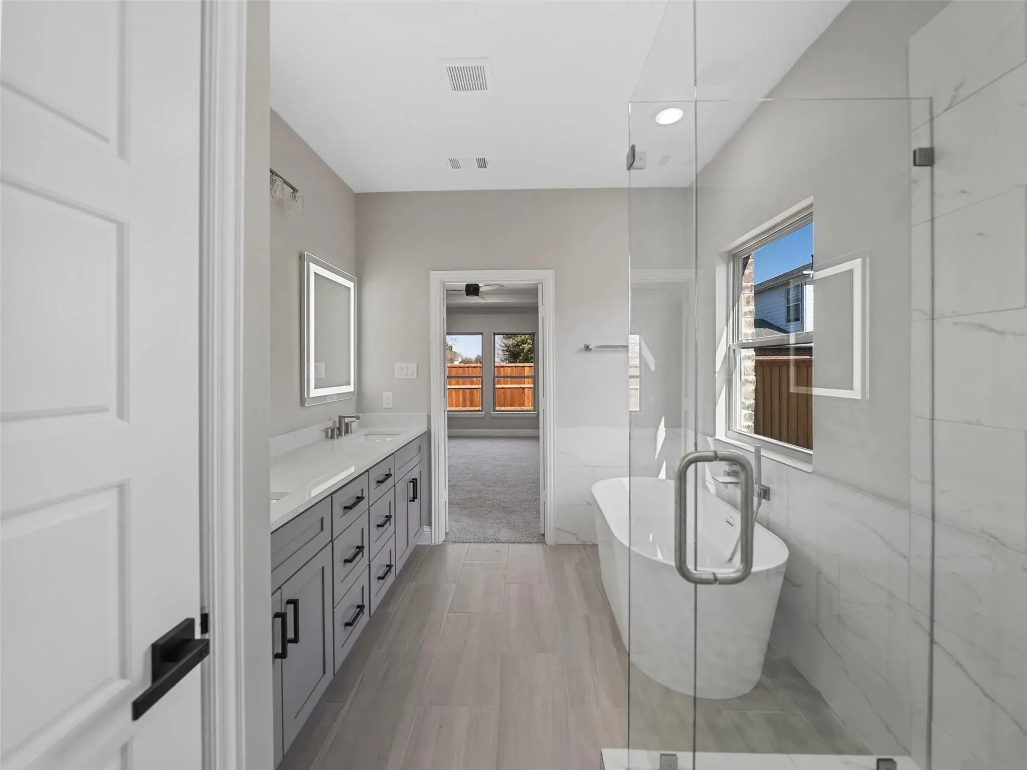 Modern bathroom featuring a freestanding soaking tub, a frameless glass shower enclosure, a dual vanity with gray cabinetry, and light-tone wood-finish flooring