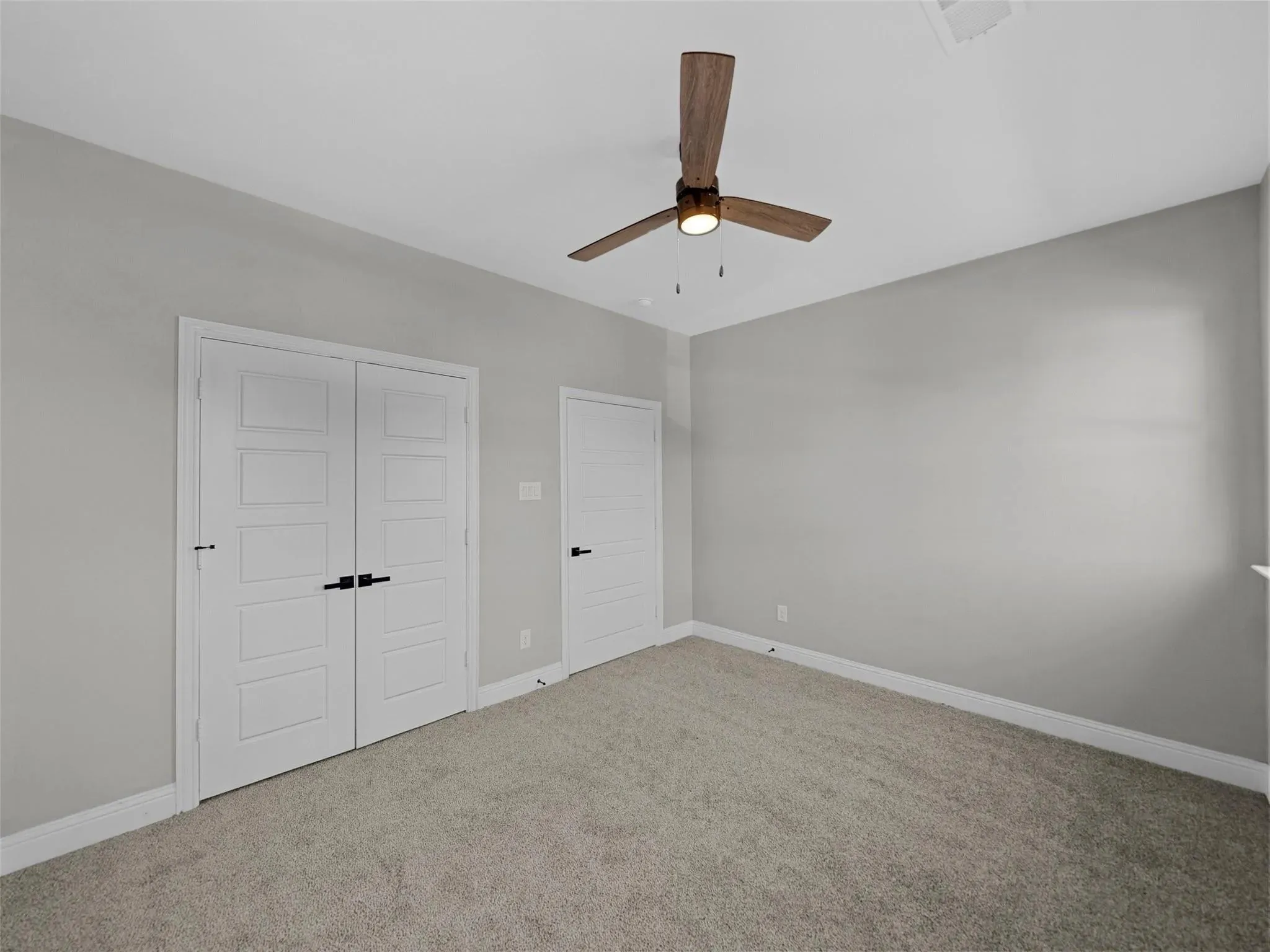 Room featuring light gray wall paint, gray carpet, a wood-finish ceiling fan with integrated lighting, white baseboards, and two white panel doors with matte black hardware