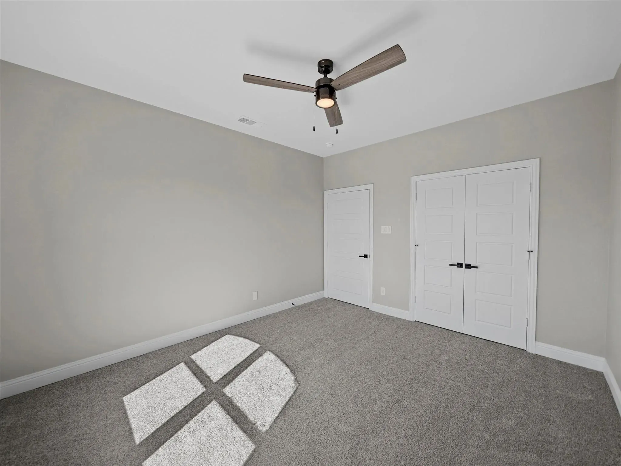 Spacious room with light gray walls, gray carpeting, a contemporary ceiling fan with light fixture, and two white paneled doors with black hardware